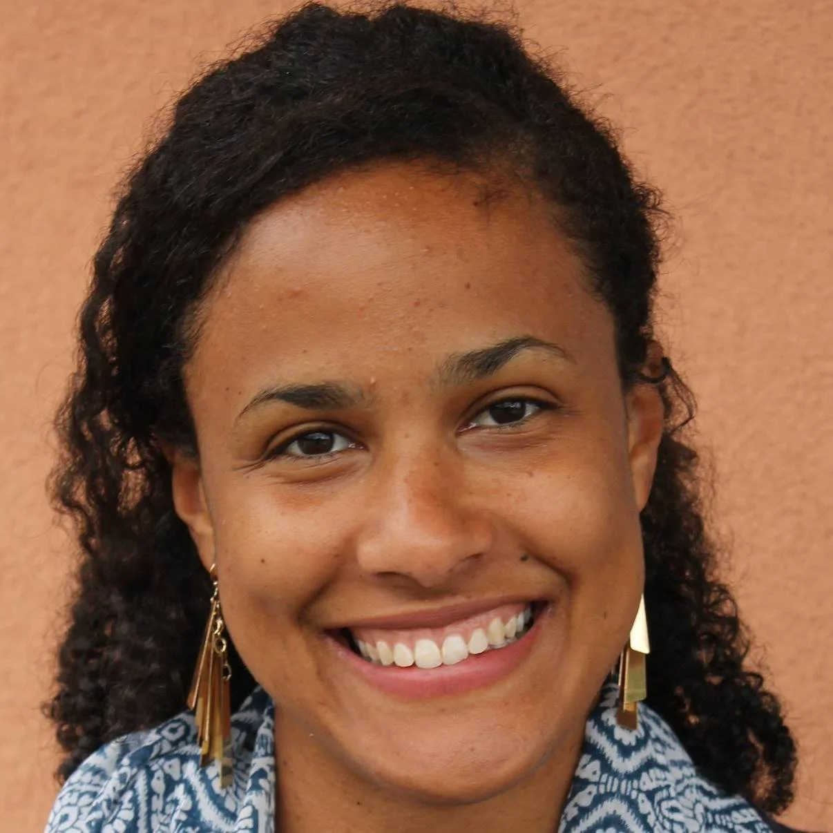 Smiling person with curly hair wearing geometric earrings and a patterned scarf against a beige background.