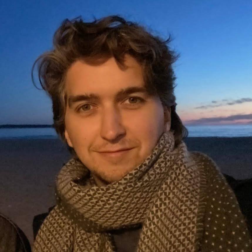 Person wearing a patterned scarf standing outdoors at sunset with a beach and ocean in the background.