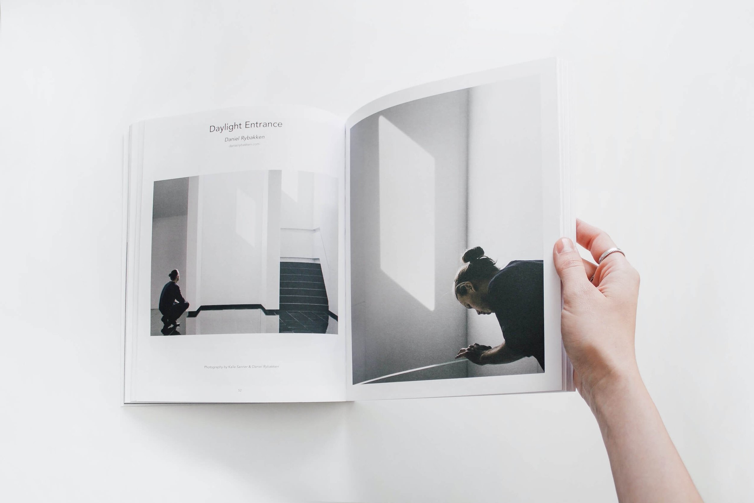 An open photo book showing black and white images of a person in a minimalist space, with one photo of a woman crouching near a staircase and another of a woman looking down in a corner