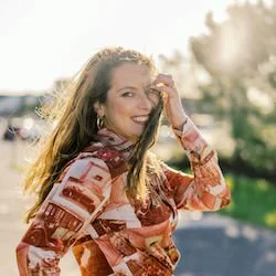 A woman smiling outdoors with sunlight behind her, wearing a patterned blouse and hoop earrings.