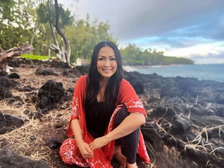 Woman sitting on rocky shoreline near the ocean, smiling, with trees and cloudy sky in the background.