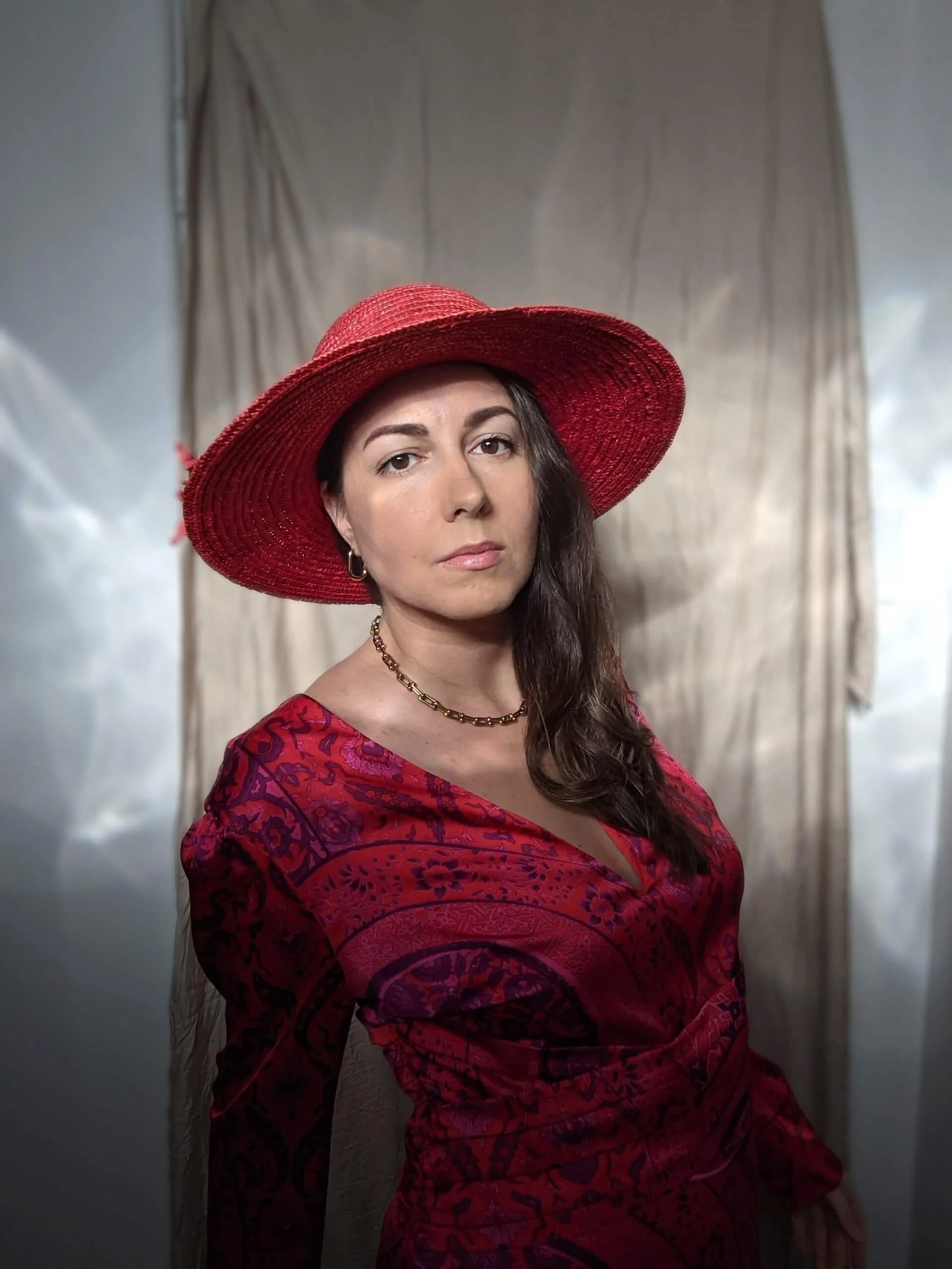 A woman with long brown hair wearing a red hat and a matching red dress with floral patterns, standing in front of a neutral background with draped fabric.