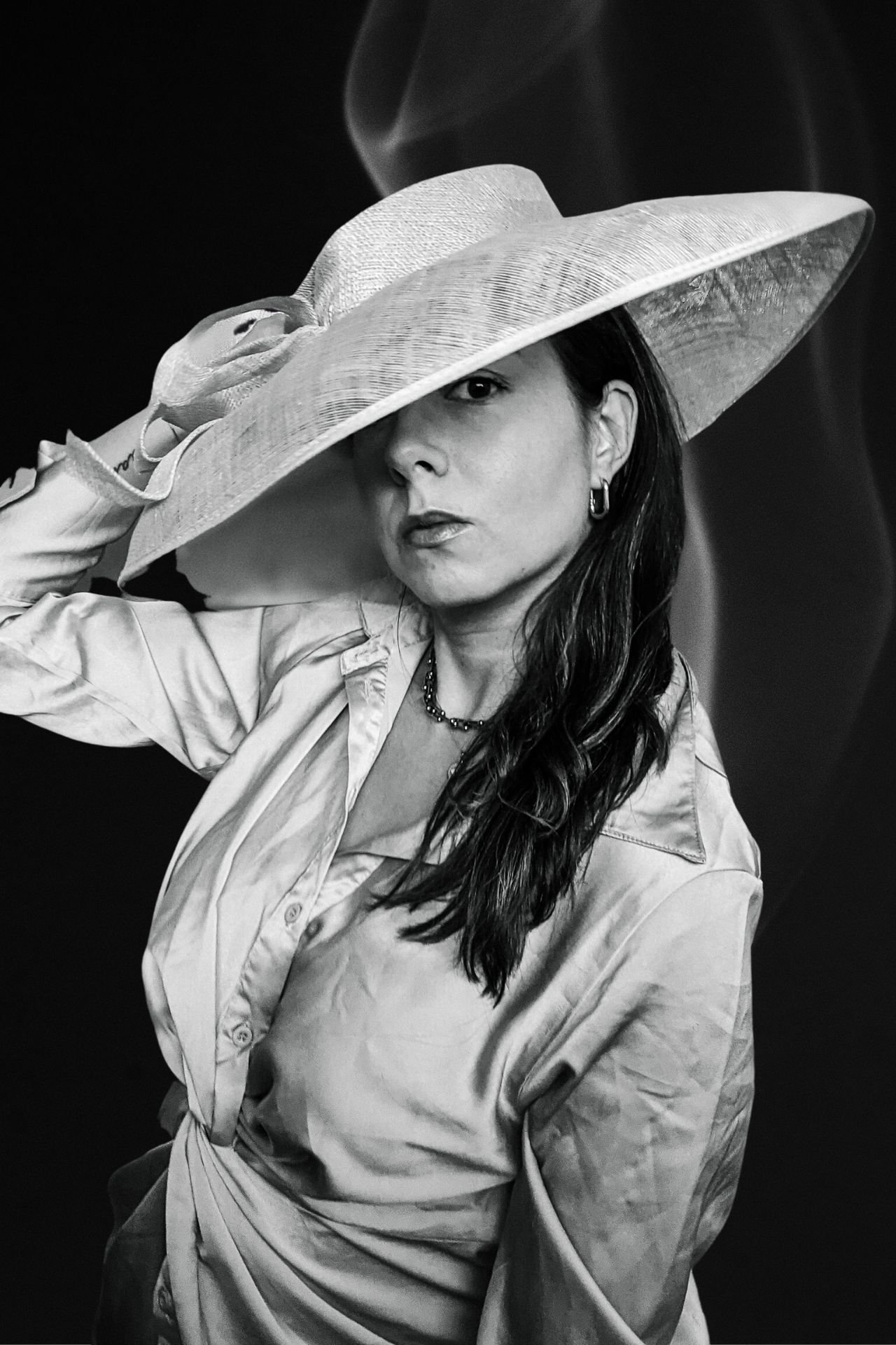 A woman wearing a wide-brimmed hat, a button-up shirt, and jewelry, posing against a dark background.