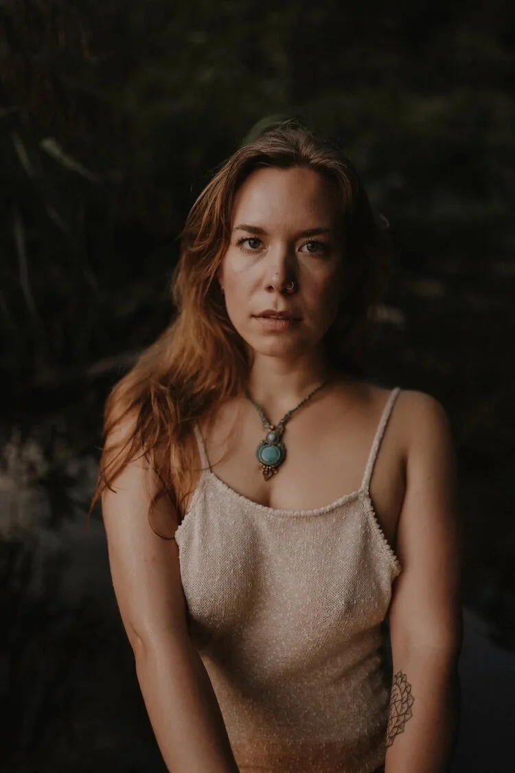 A young woman with long red hair, wearing a beige sleeveless top and a turquoise pendant necklace, looking directly at the camera with a neutral expression, dark background.