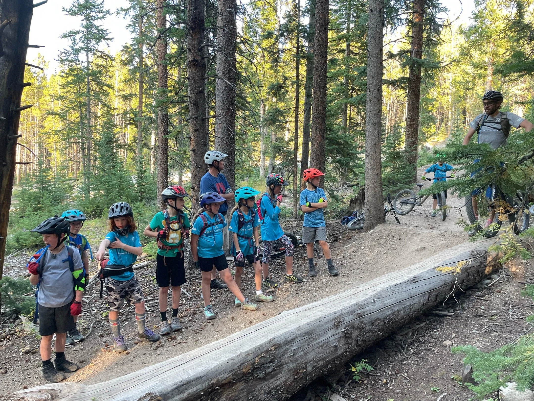A group of children wearing helmets and bikes are standing on a forest trail, listening to an adult male instructing them as he stands next to a fallen tree on the trail.
