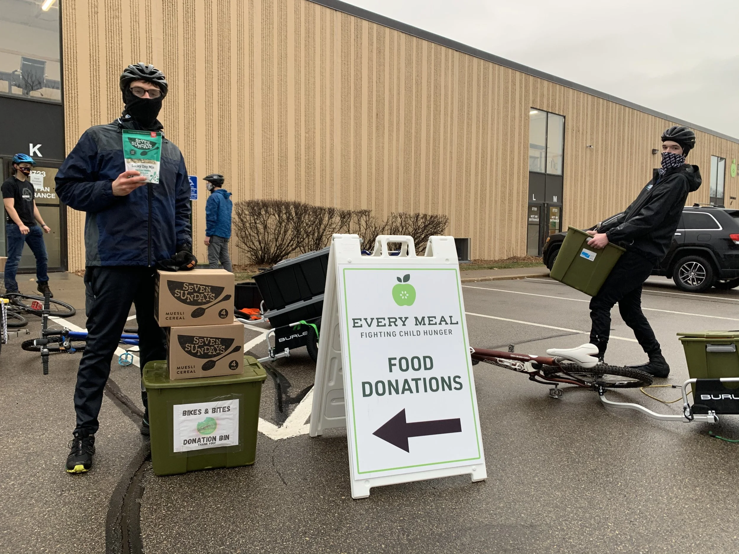 Two people with bicycles and helmets standing near a donation bin, holding food items, at a food donation event outside a building. One person holds a package of cereal, and a sign indicates this is a food donation drive to fight child hunger.