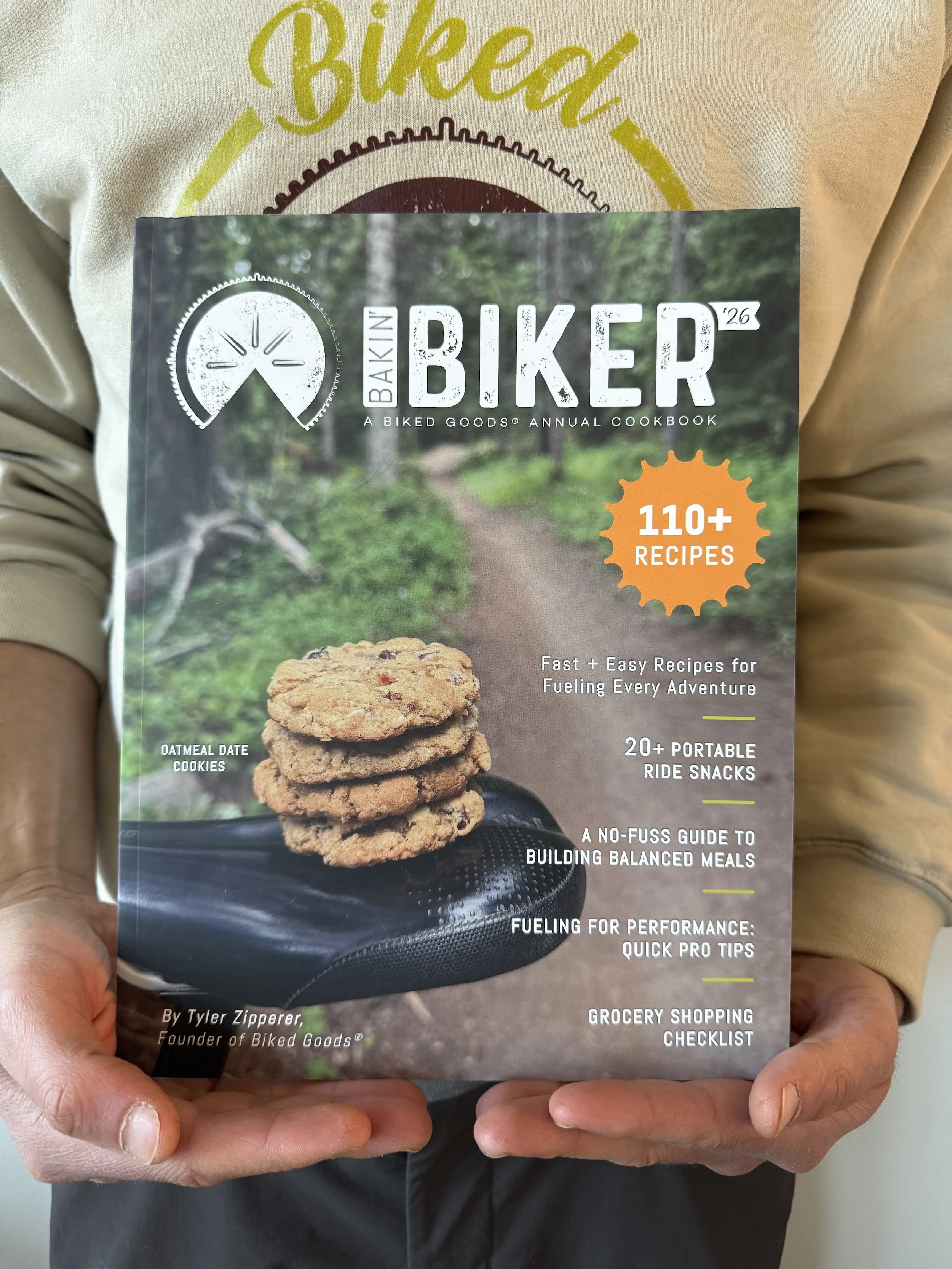 Person holding a cookbook titled 'Baking Biker,' featuring a stack of oatmeal date cookies on the cover, with a nature trail background.