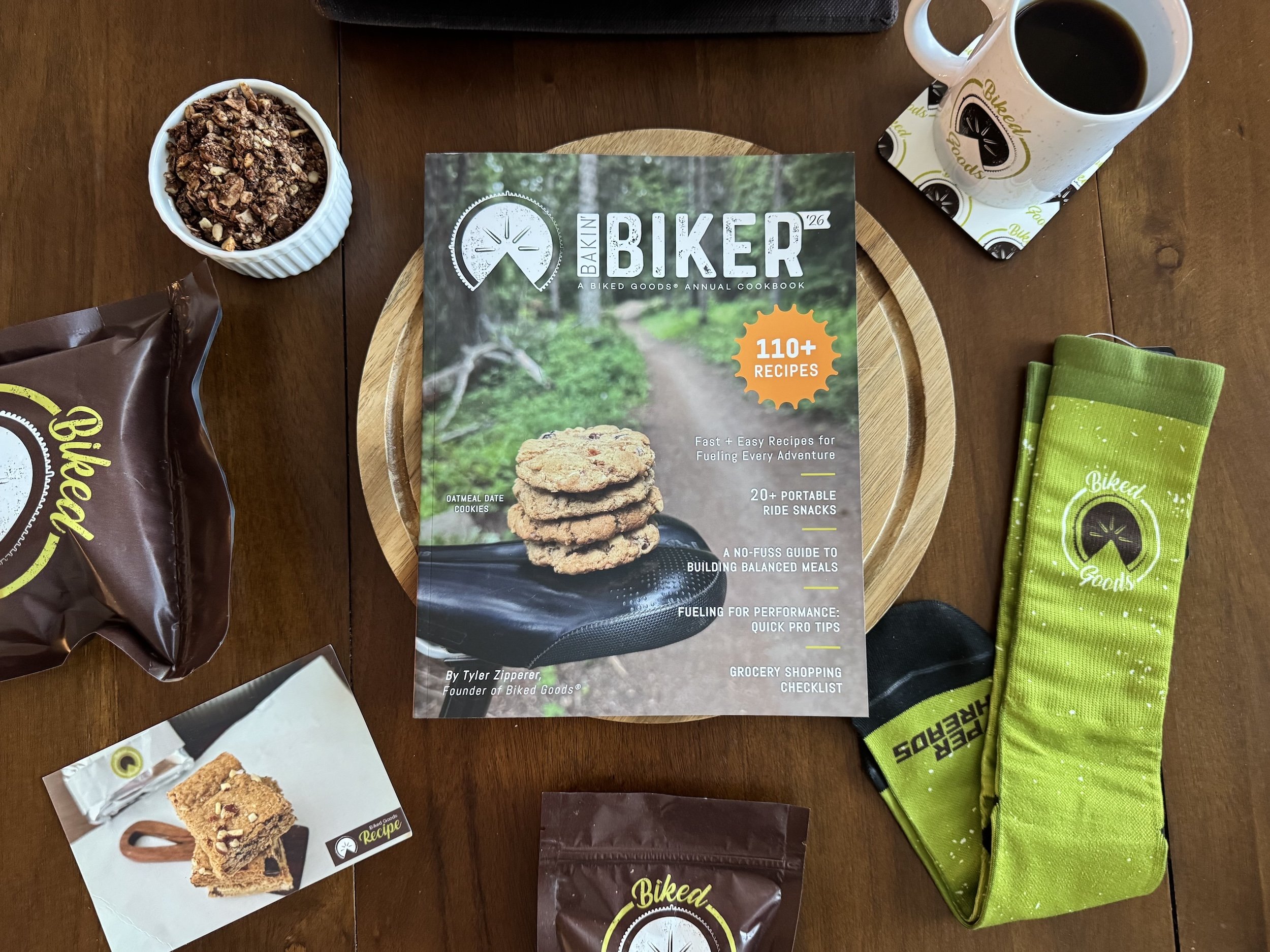 A cookbook titled 'Baker Biker' surrounded by snacks and items on a wooden table. Includes a bag of Biked Goods cookies, a cup of black coffee, a packet of granola, a small photo of baked oatmeal bars, and a green Biked Goods towel.