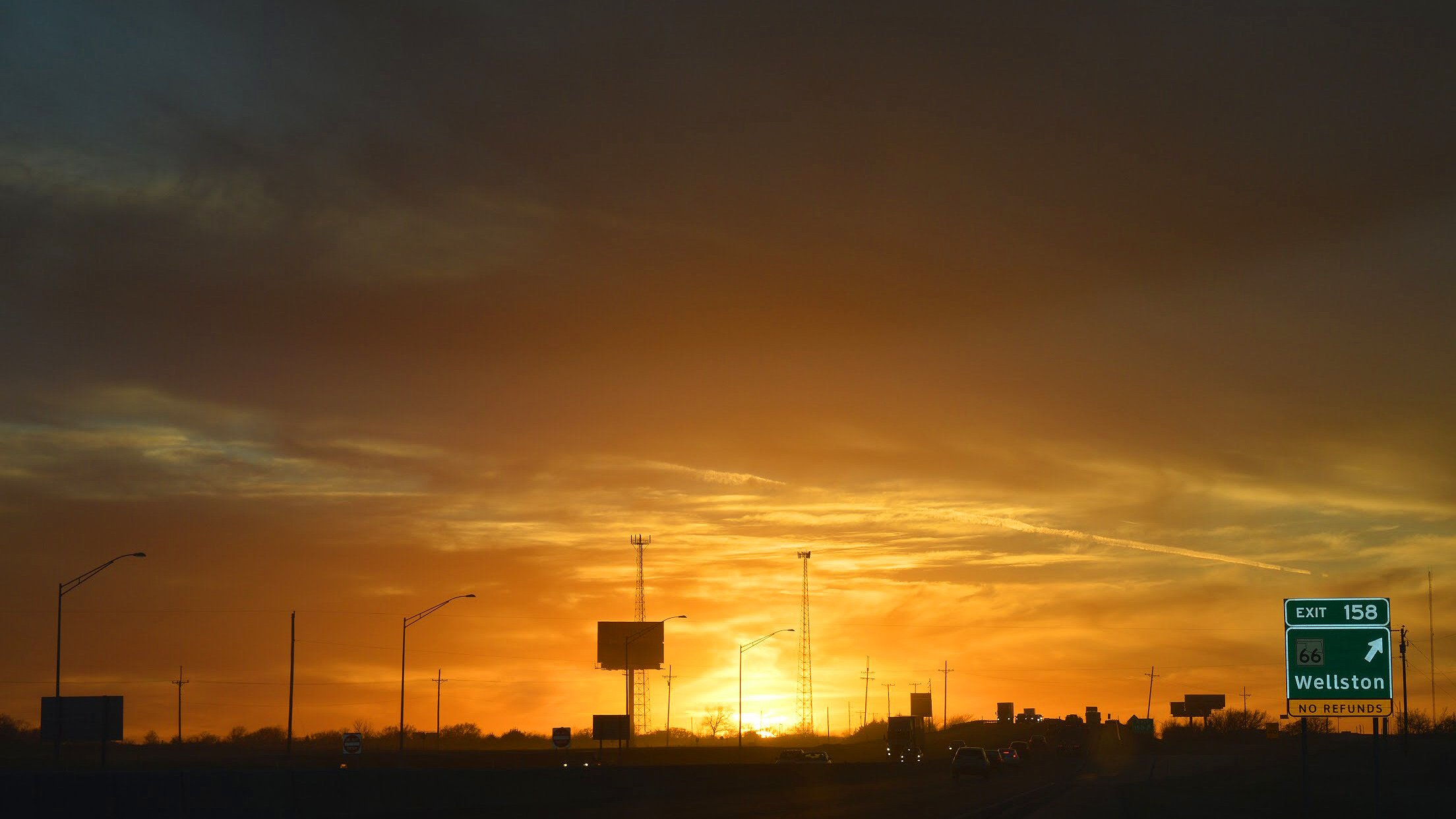 Sunset over a highway with exit sign for Wellston, tall communication towers, streetlights, and cars.