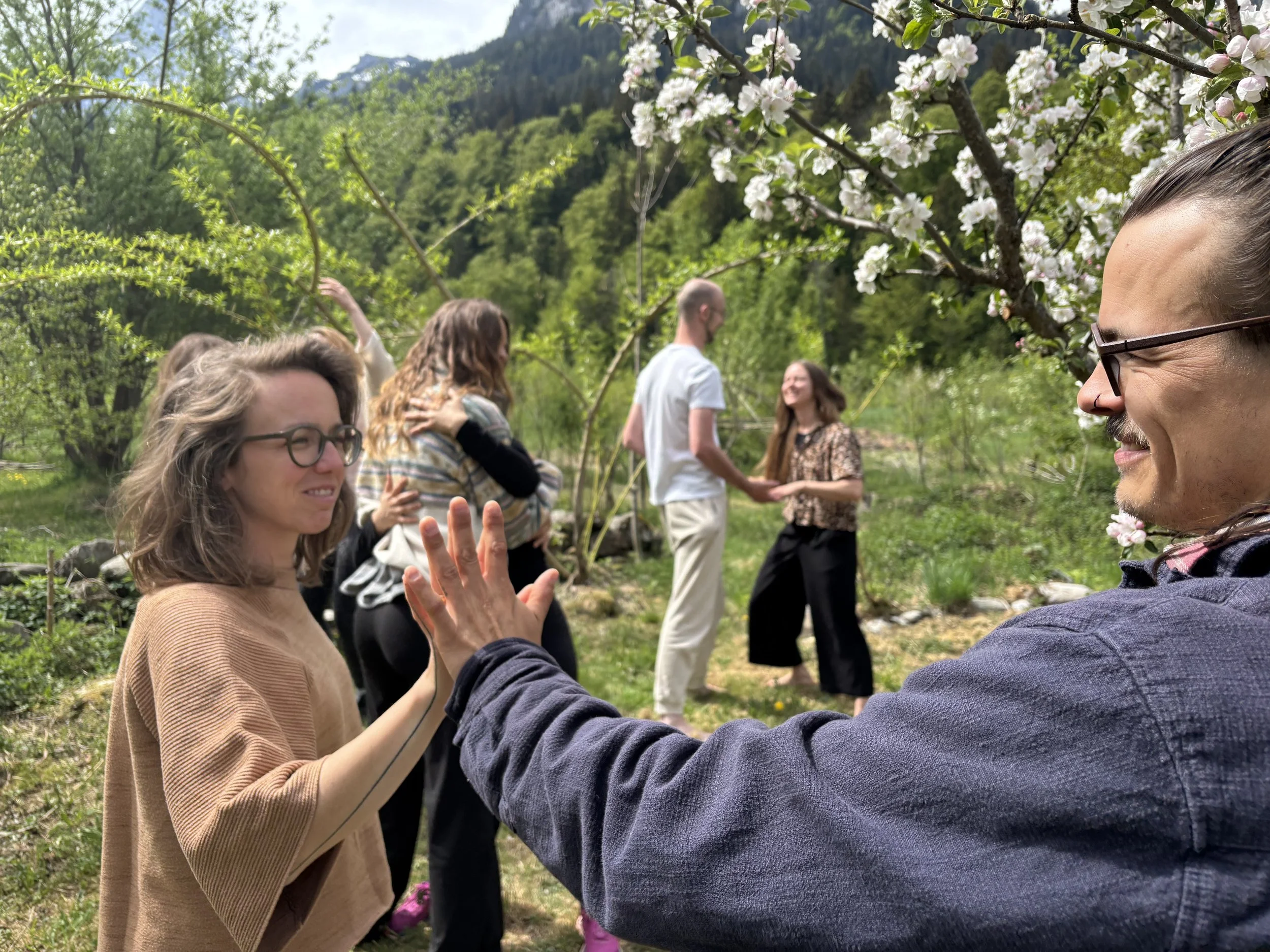 Fünf Menschen, die in einem Garten mit blühenden Bäumen und grünen Hügeln im Hintergrund stehen, scheinen sich zu umarmen und zu begrüßen, während sie fröhlich sprechen.