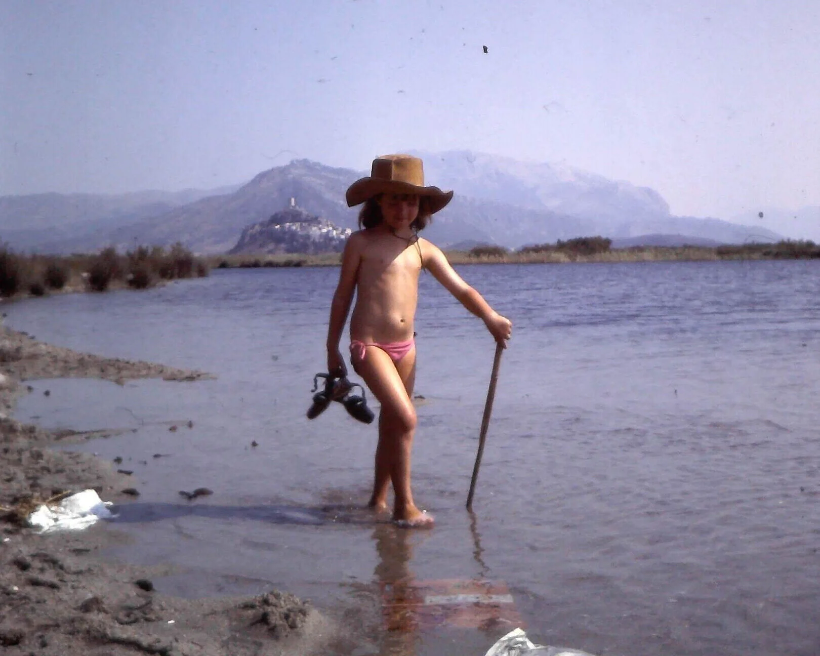 Un enfant en maillot de bain rose, portant un grand chapeau en paille, marche dans une rivière avec une canne dans la main, fond montagneux et nature environnante. C'est la psychologue quand elle était enfant.