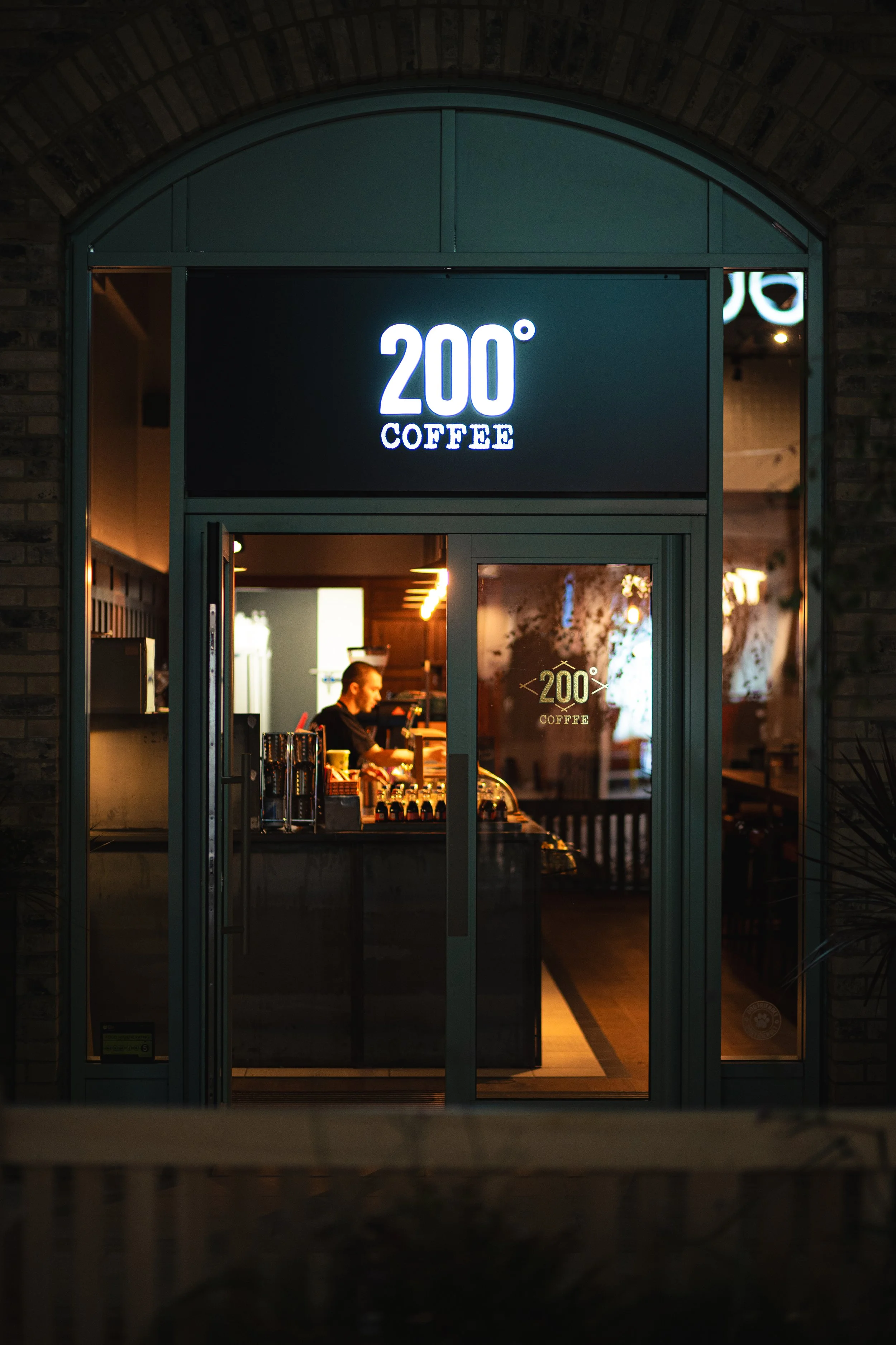 Night view of a coffee shop called 200° Coffee, seen through glass doors with a person working behind the counter inside.
