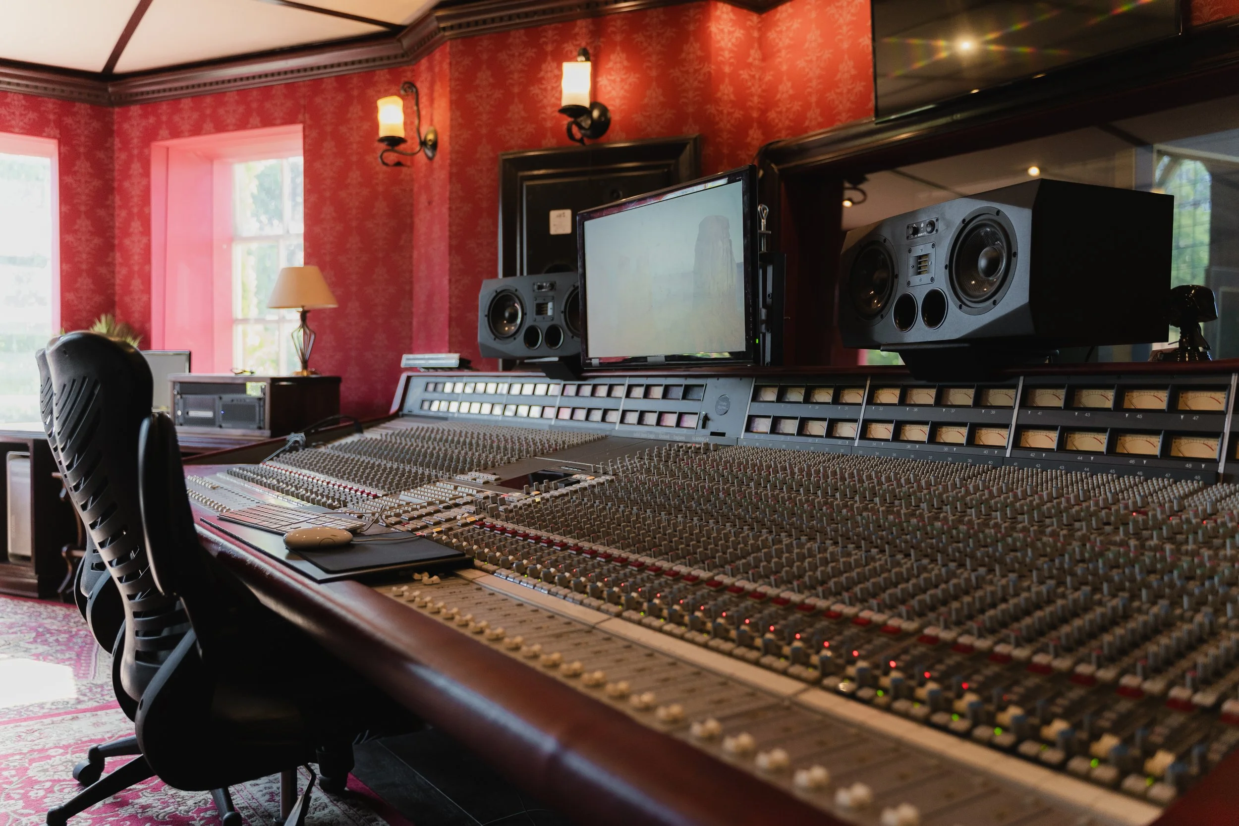 Professional recording studio with a large mixing console, audio speakers, a computer monitor, and a red textured wall in the background.