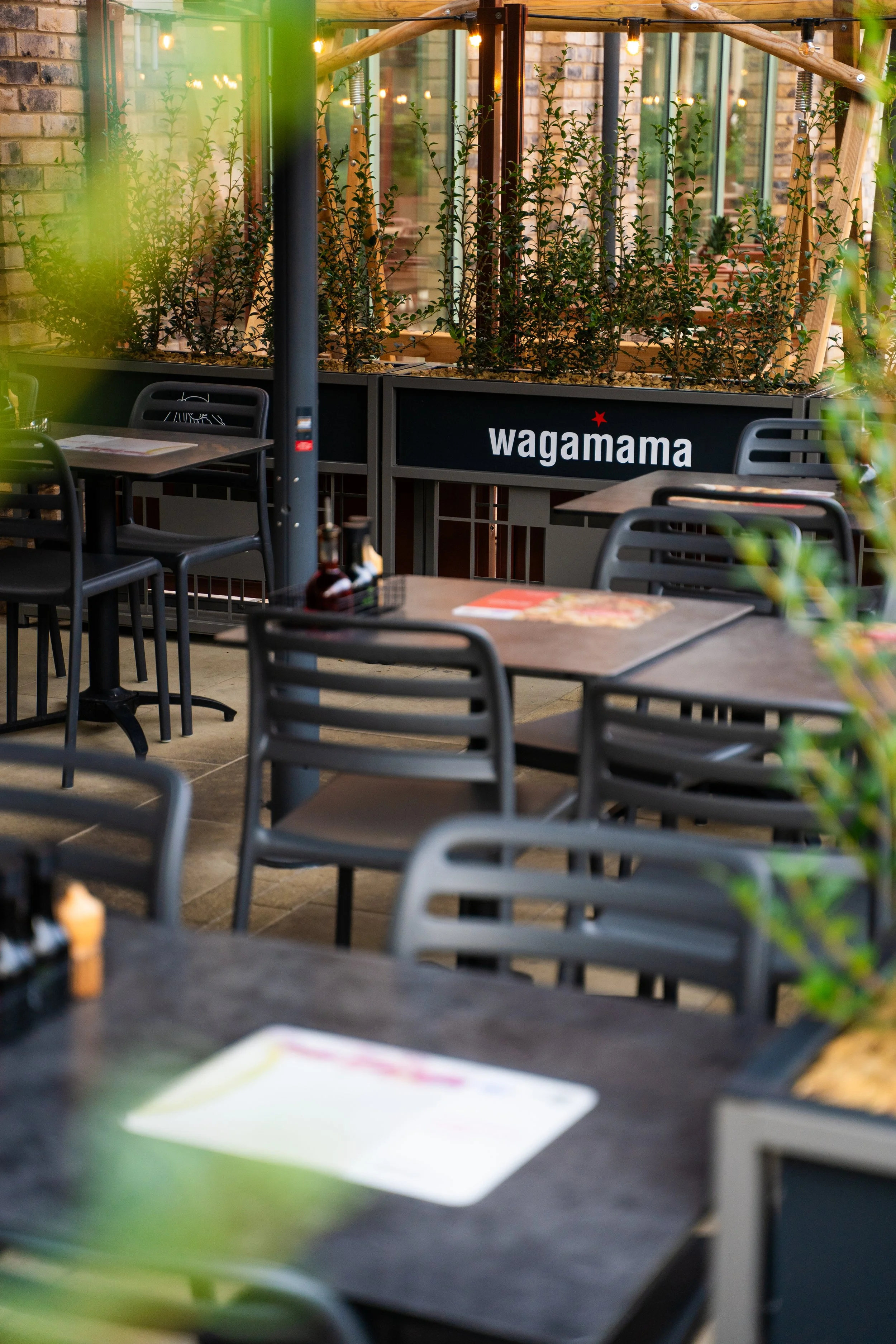 Empty outdoor patio seating area with black tables and chairs, green plants, and a black sign with white text that reads "wagamama".