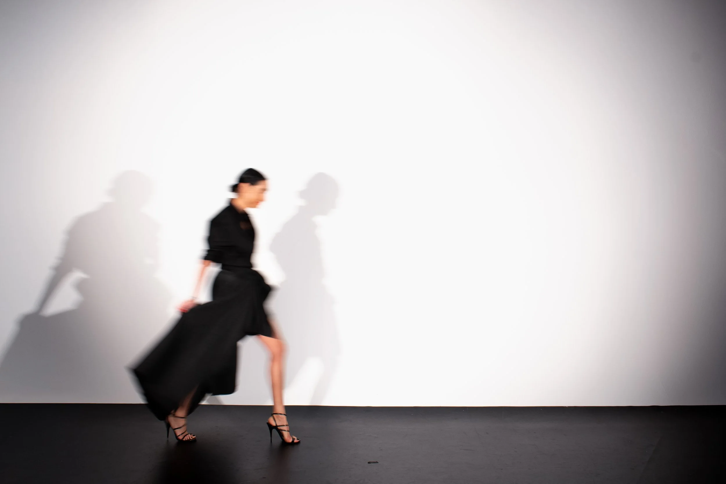 A blurred woman in a black dress and high heels walking against a plain white wall, with her shadow projected on the wall.