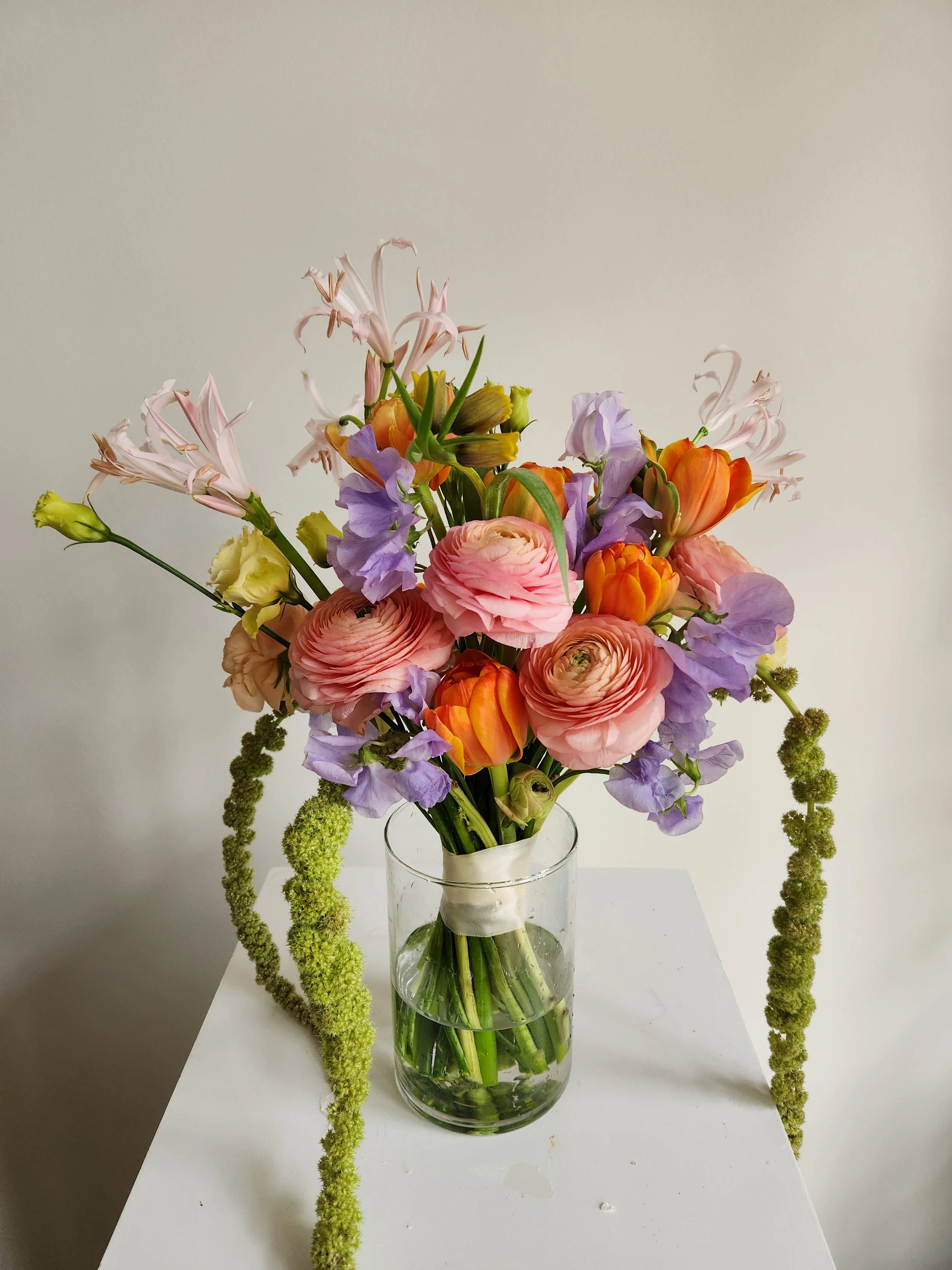 A bouquet of mixed flowers including pink ranunculus, purple sweet peas, orange tulips, and pale yellow lisianthus in a clear glass vase on a white surface against a plain white background.