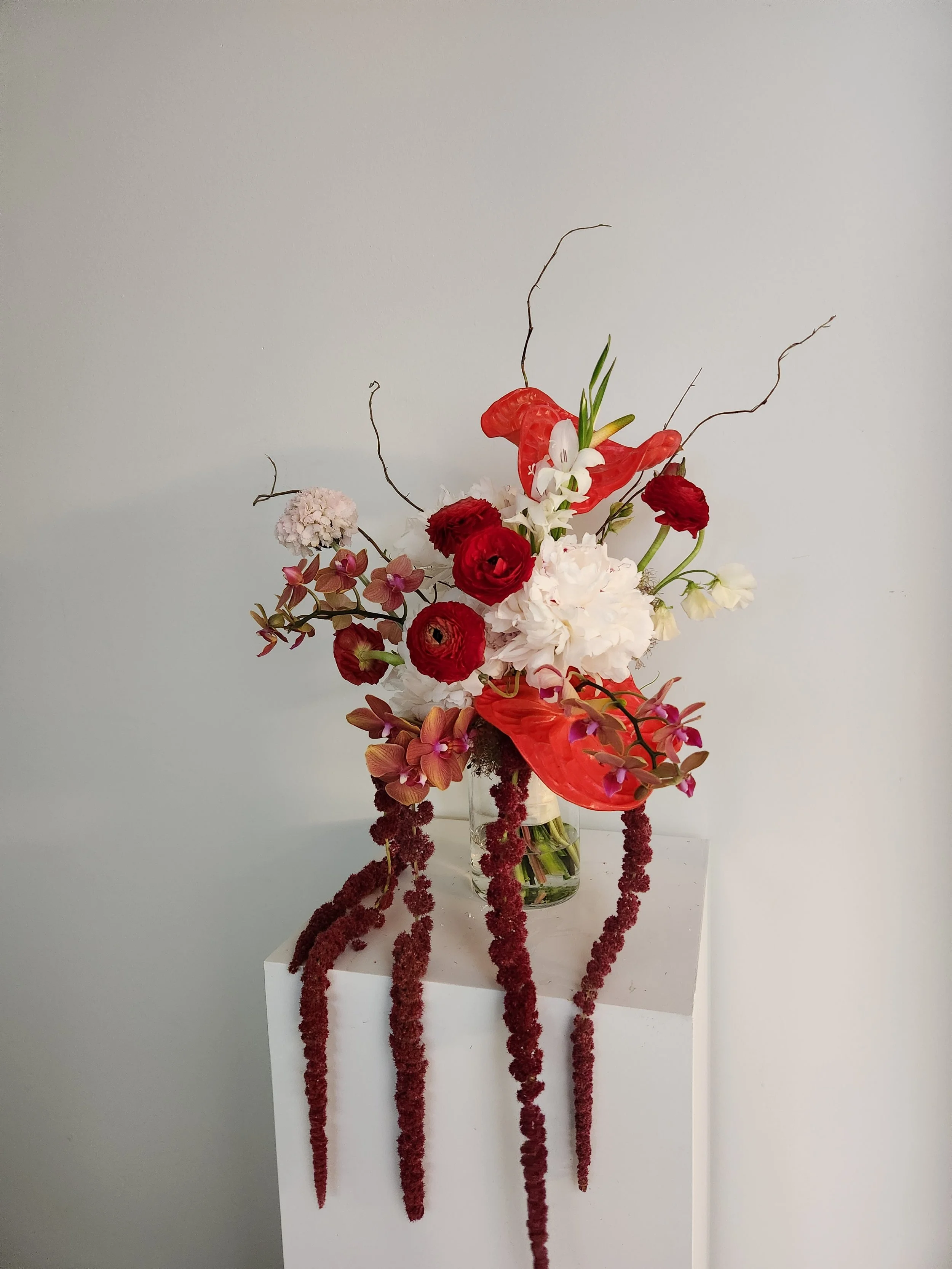 Elegant wedding floral arrangement with red, white, pink, and purple flowers in a clear glass vase on a white pedestal against a pale gray background.