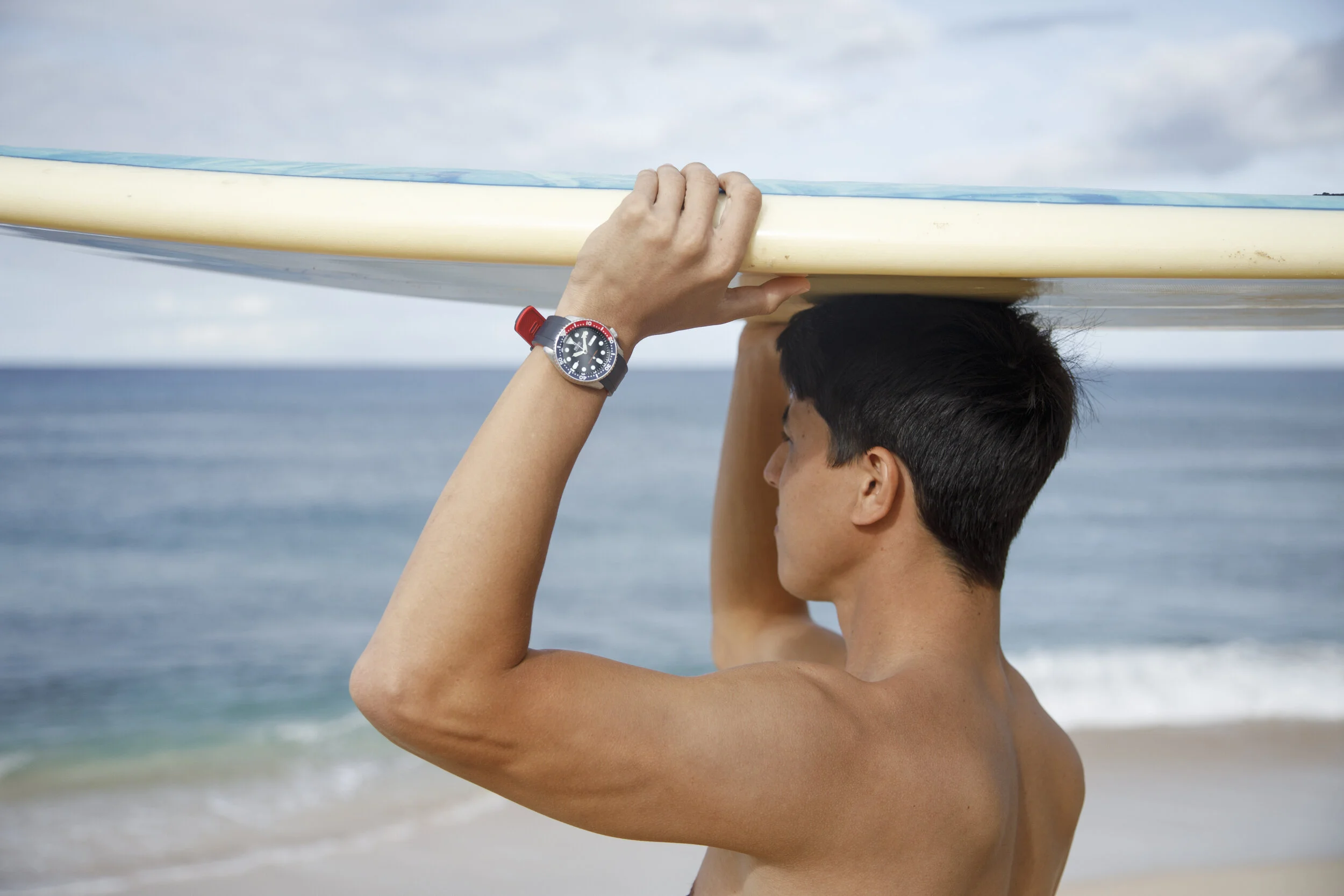 Man carrying wearing a Seiko watch, carrying a surfboard on his head at the beach, ocean in the background.