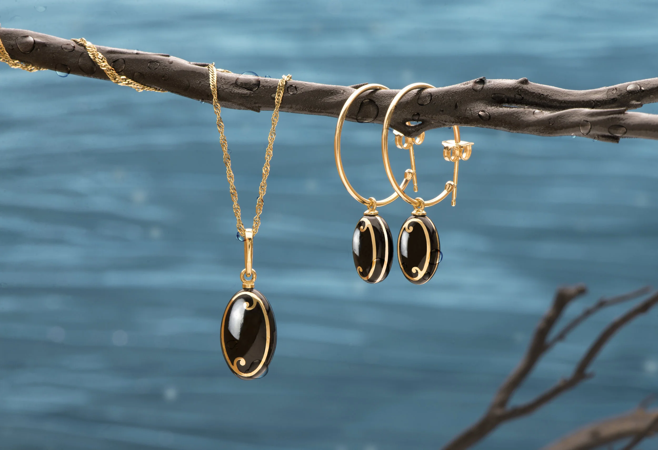 Gold Maui Divers Jewelry Endless Ocean Collection including a necklace and earrings with black and gold oval pendants, hanging on a water-drenched branch over a body of water.