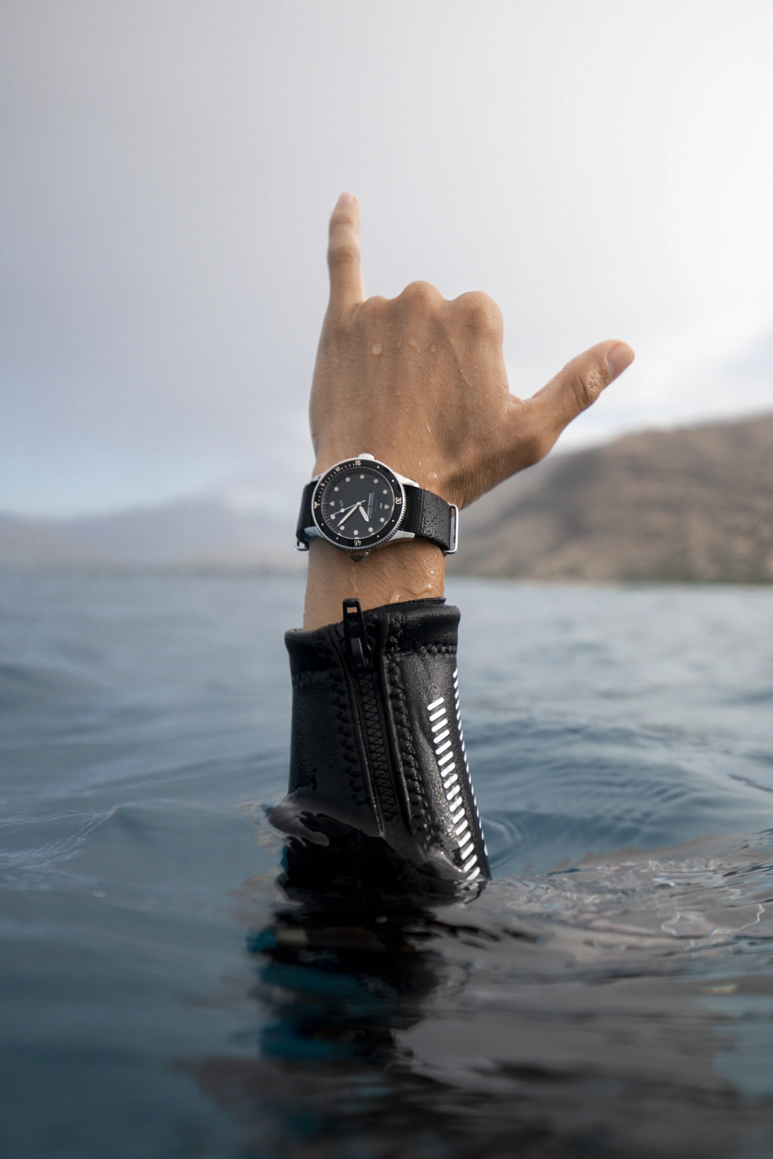 A person's arm is raised above water, making a hand gesture, wearing a black watch and a black water-resistant jacket. The background features a cloudy sky and a distant shoreline.