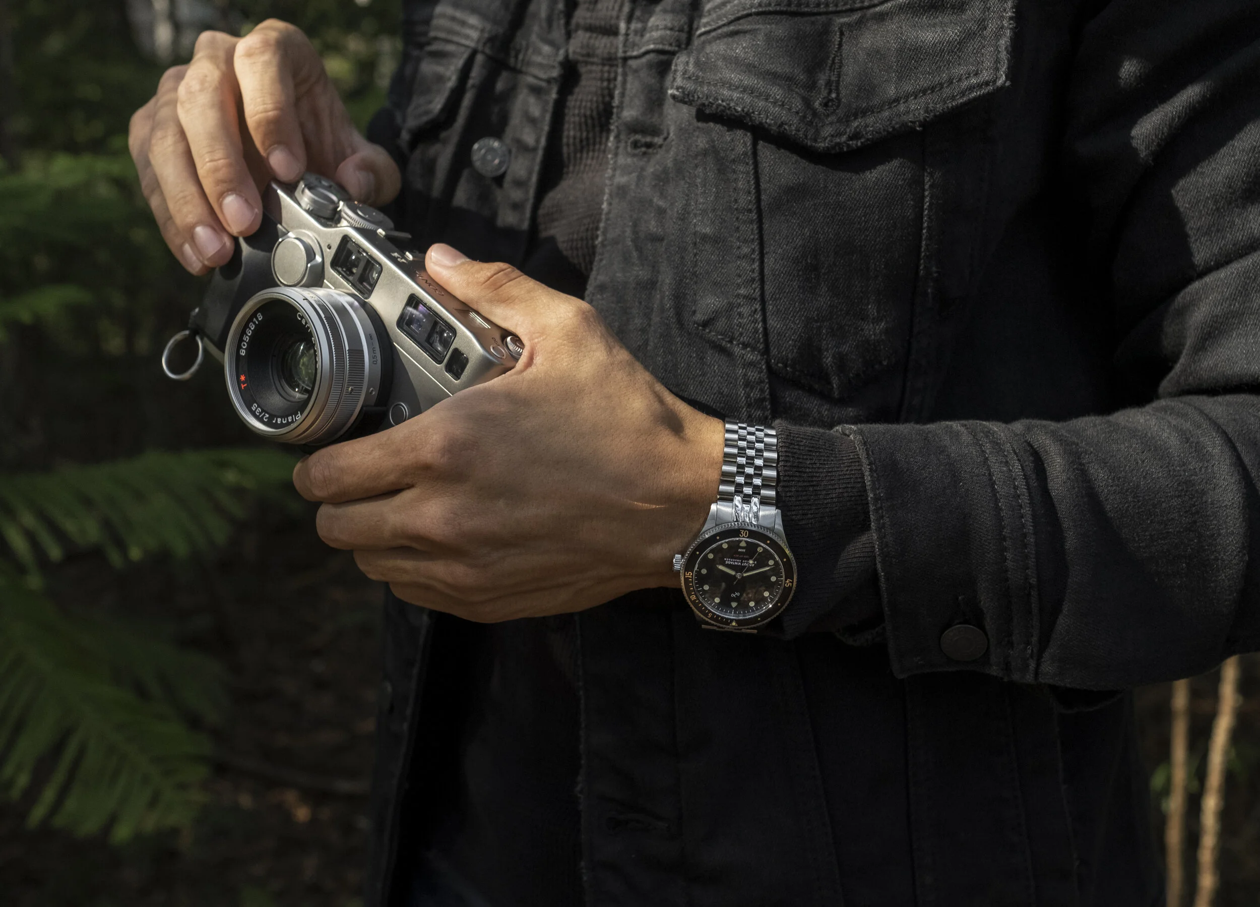 Person wearing a black jacket and watch holding a vintage-style camera outdoors.