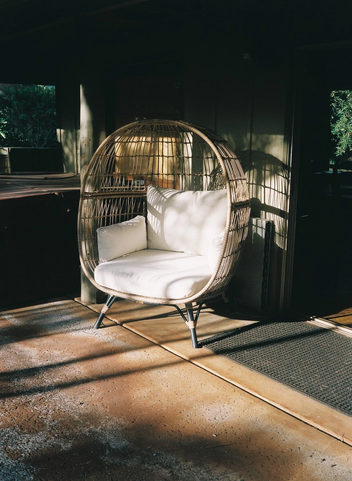 A modern wicker egg-shaped chair with white cushions placed inside a dimly lit room, sunlight casting shadows on the floor.