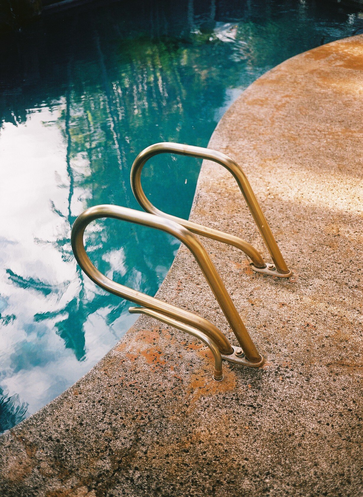 Metal pool ladder beside a swimming pool, with water reflecting the sky and nearby trees.