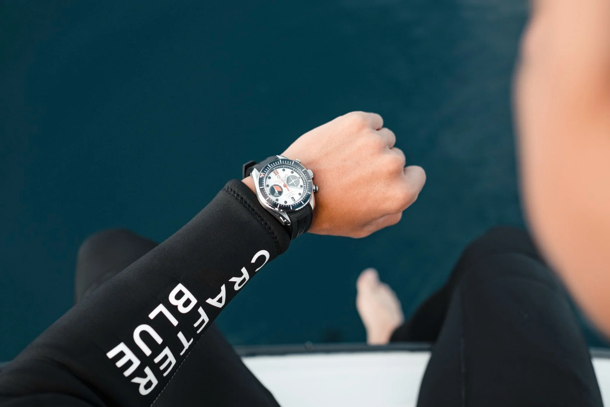 A person wearing a black wetsuit with white lettering on the sleeve, looking at a Crafter Blue wristwatch while standing near the water.