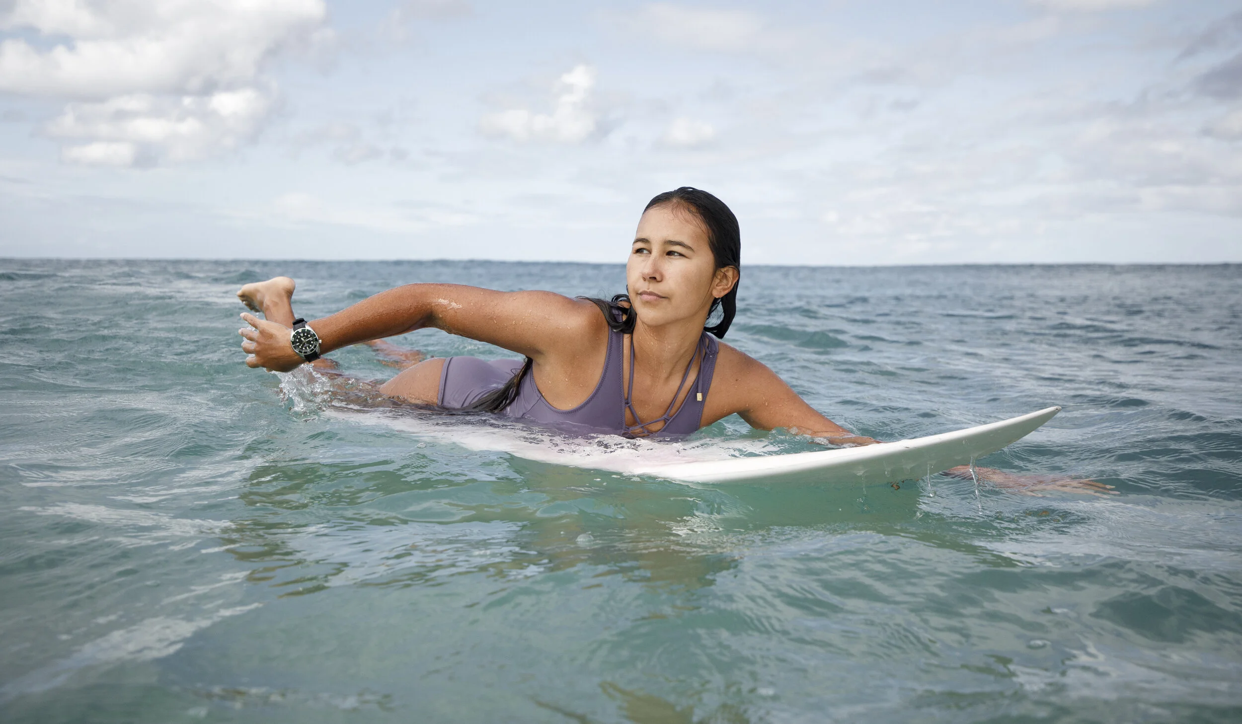 Woman in a purple swimsuit paddling on a white surfboard in the ocean under cloudy sky.