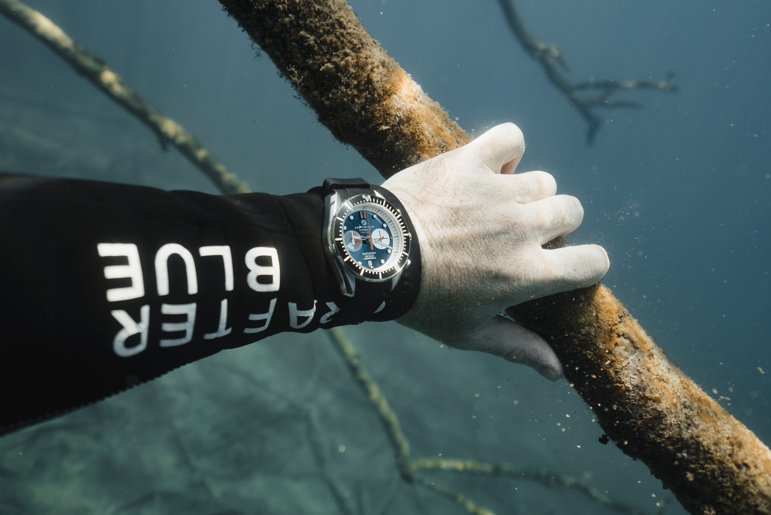 A person's hand holding onto a rusty, moss-covered branch underwater, wearing a black wetsuit with white text and a black and blue Crafter Blue diving watch.