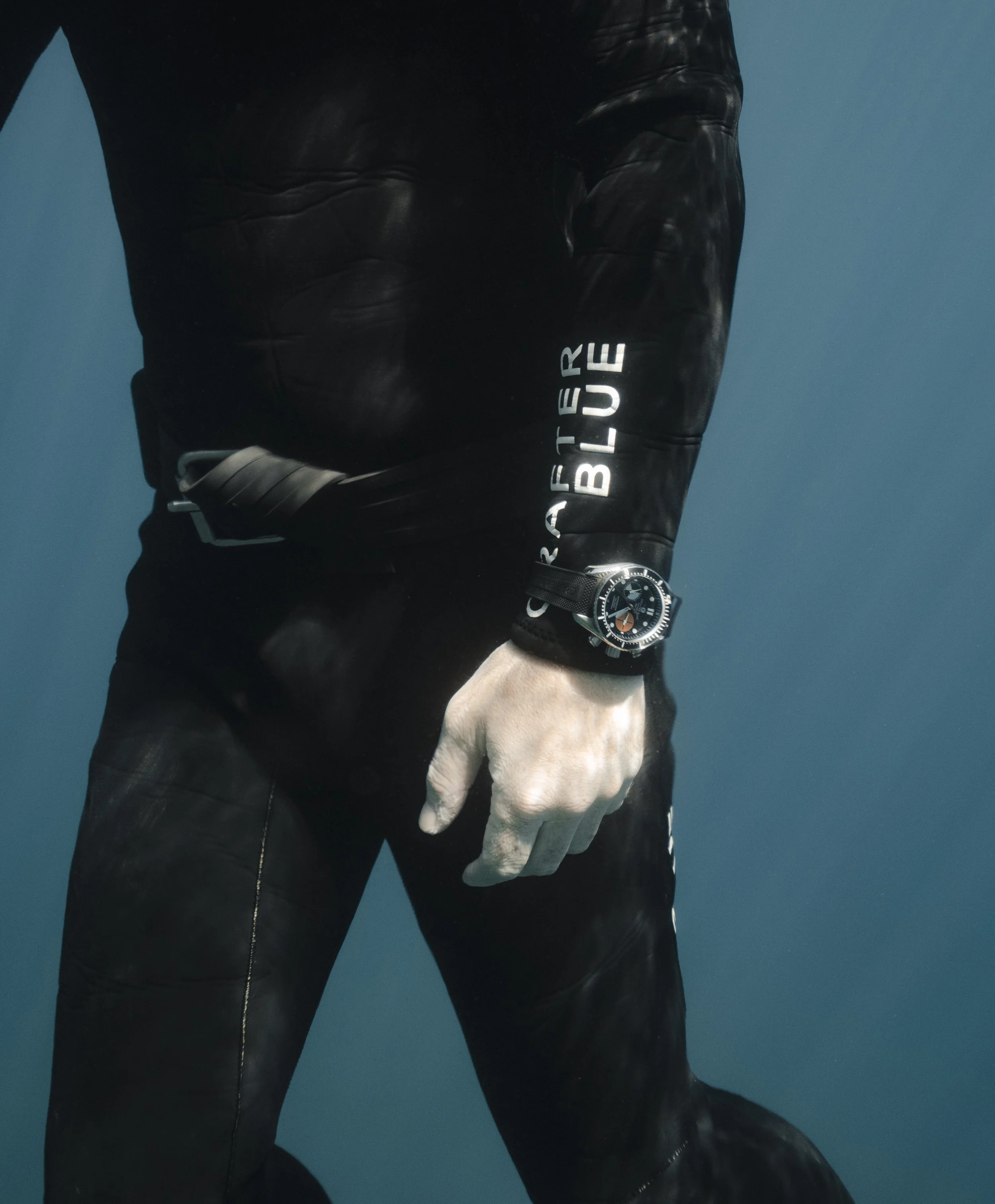 Close-up of a person's arm and hand, wearing a black leather jacket and a large dark Crafter Blue wristwatch, against a blue background.