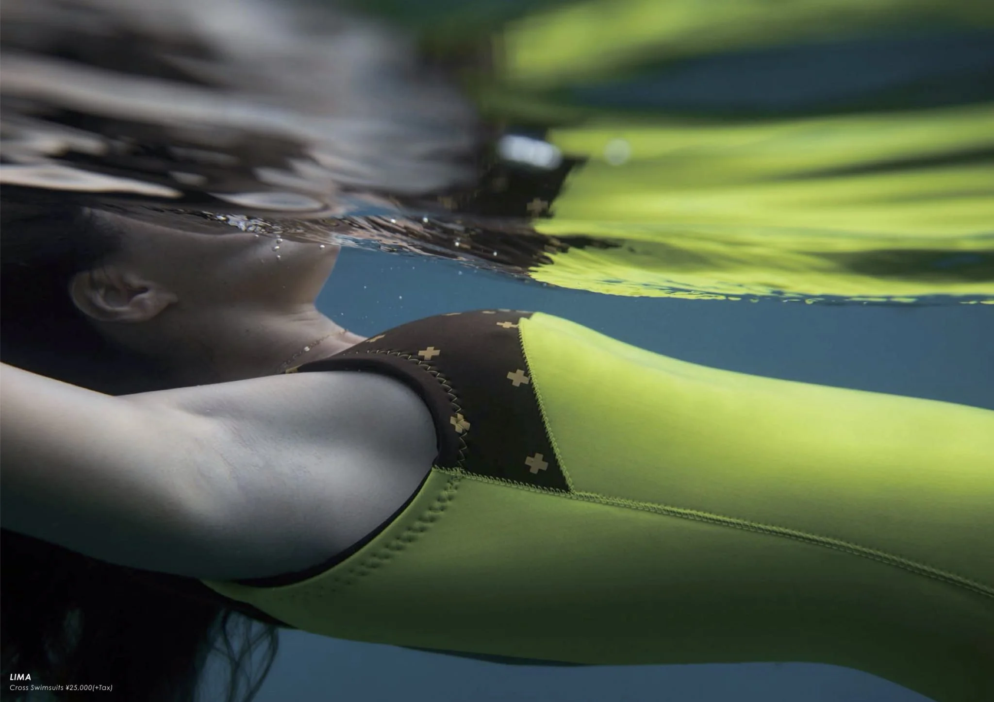 A person wearing neon yellow and black cross swimsuit is floating on water, with their face partially submerged and head turned to the side, showing their ear and part of their face from below the water surface.
