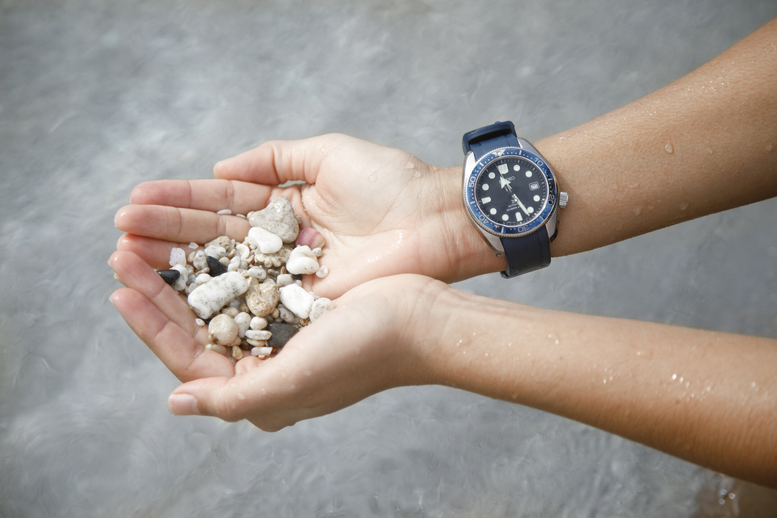 A person holding small rocks and pebbles in their hands, with water droplets on their arms, wearing a black and blue Seiko watch.