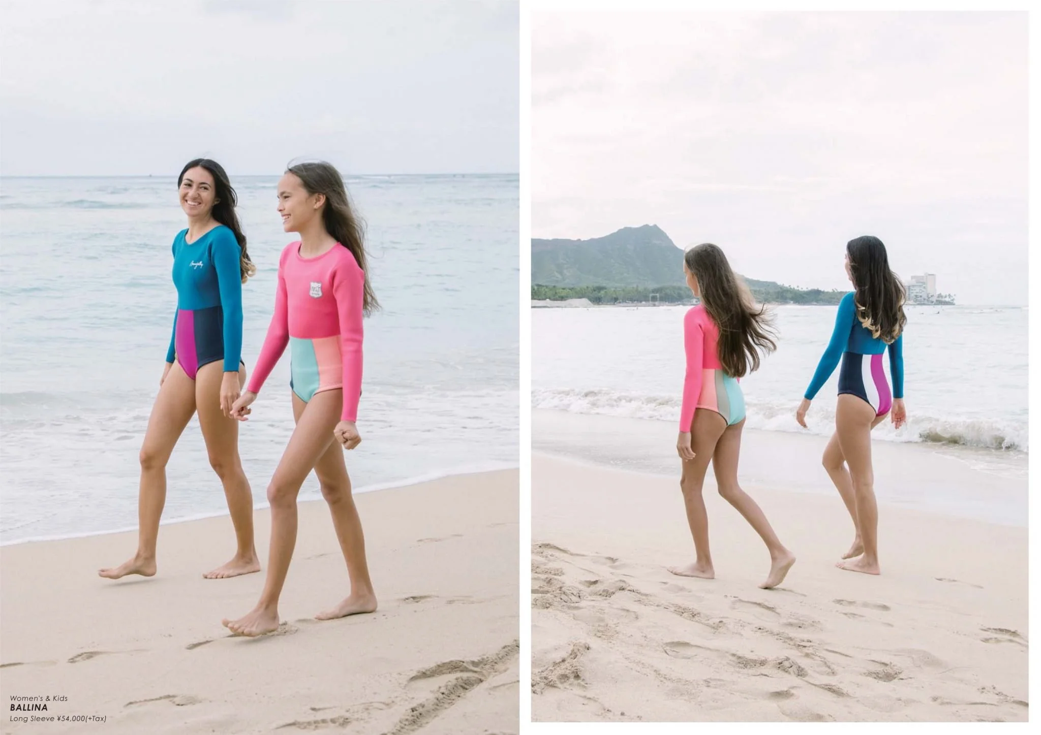 Two women, one in a blue long-sleeve swimsuit and the other in a pink long-sleeve swimsuit, walking on the beach holding hands along the shoreline with ocean and hills in the background.