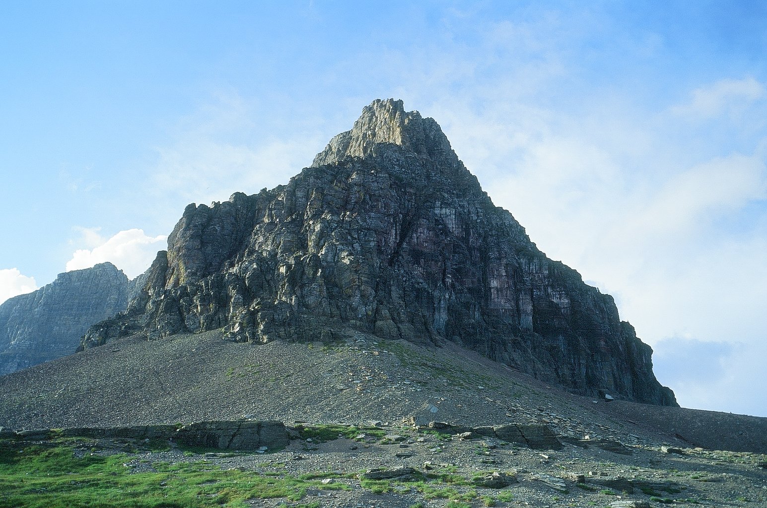 A large rocky mountain with a steep, jagged peak, green grassy foreground, and a partly cloudy sky in the background.