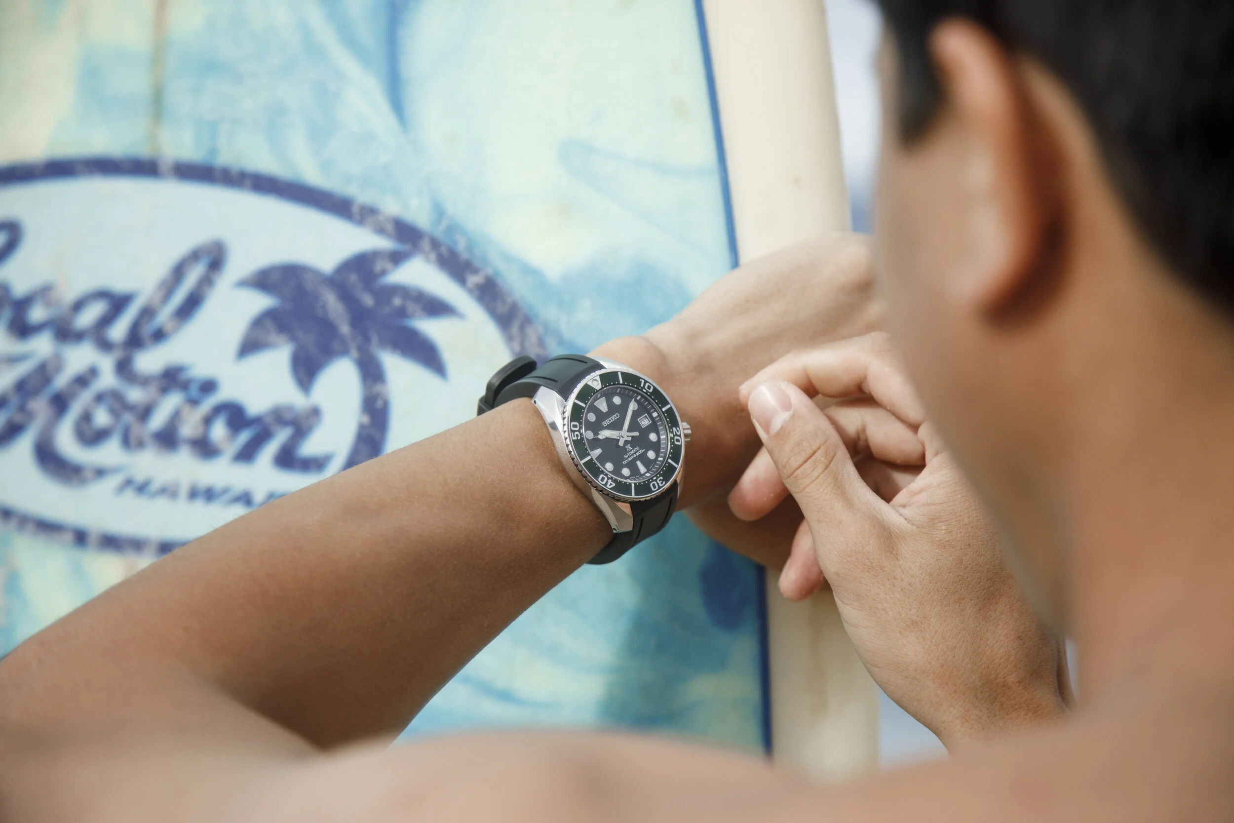 Person wearing a Seiko diving watch checking time on their wrist while standing near a surfboard with a Hawaii logo.