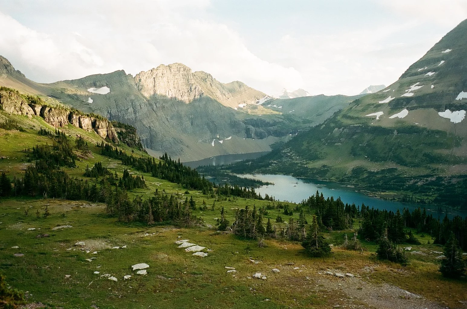 A scenic mountain landscape with a lake, green hills, trees, and partly cloudy sky.