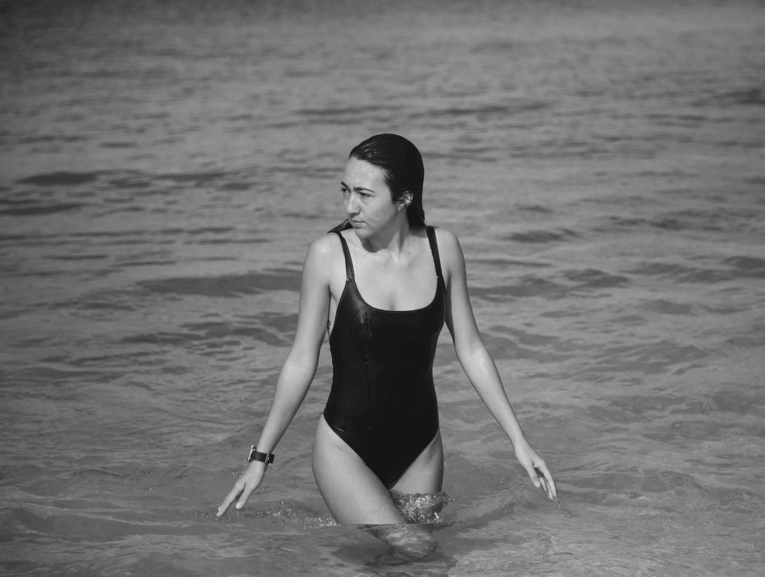 Black and white photo of a woman in a swimsuit walking out of the water at the beach, with water splashing around her.
