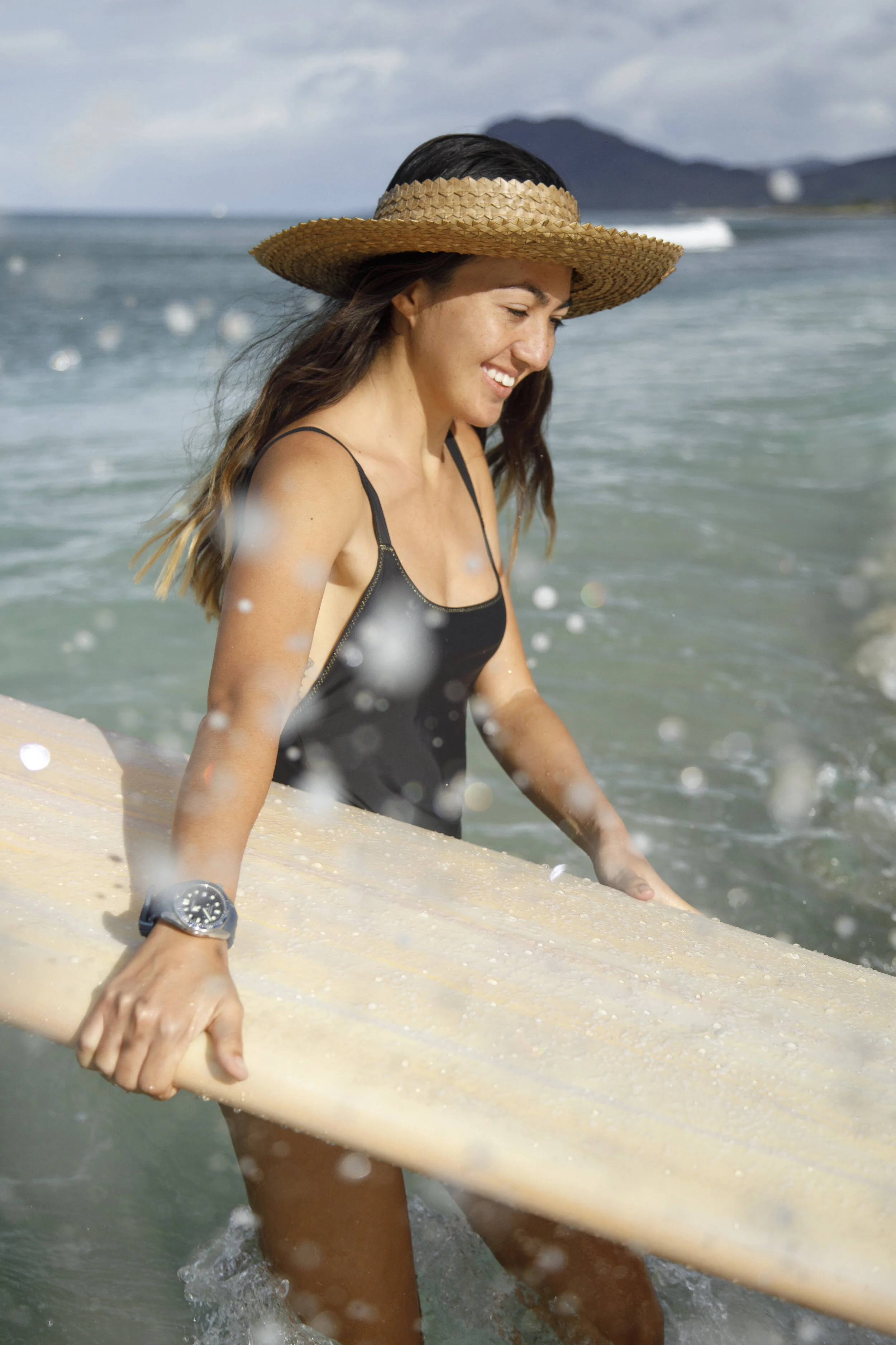 A woman smiling, wearing a straw hat, black swimsuit, and Seiko watch, carrying a surfboard into the ocean at a beach.