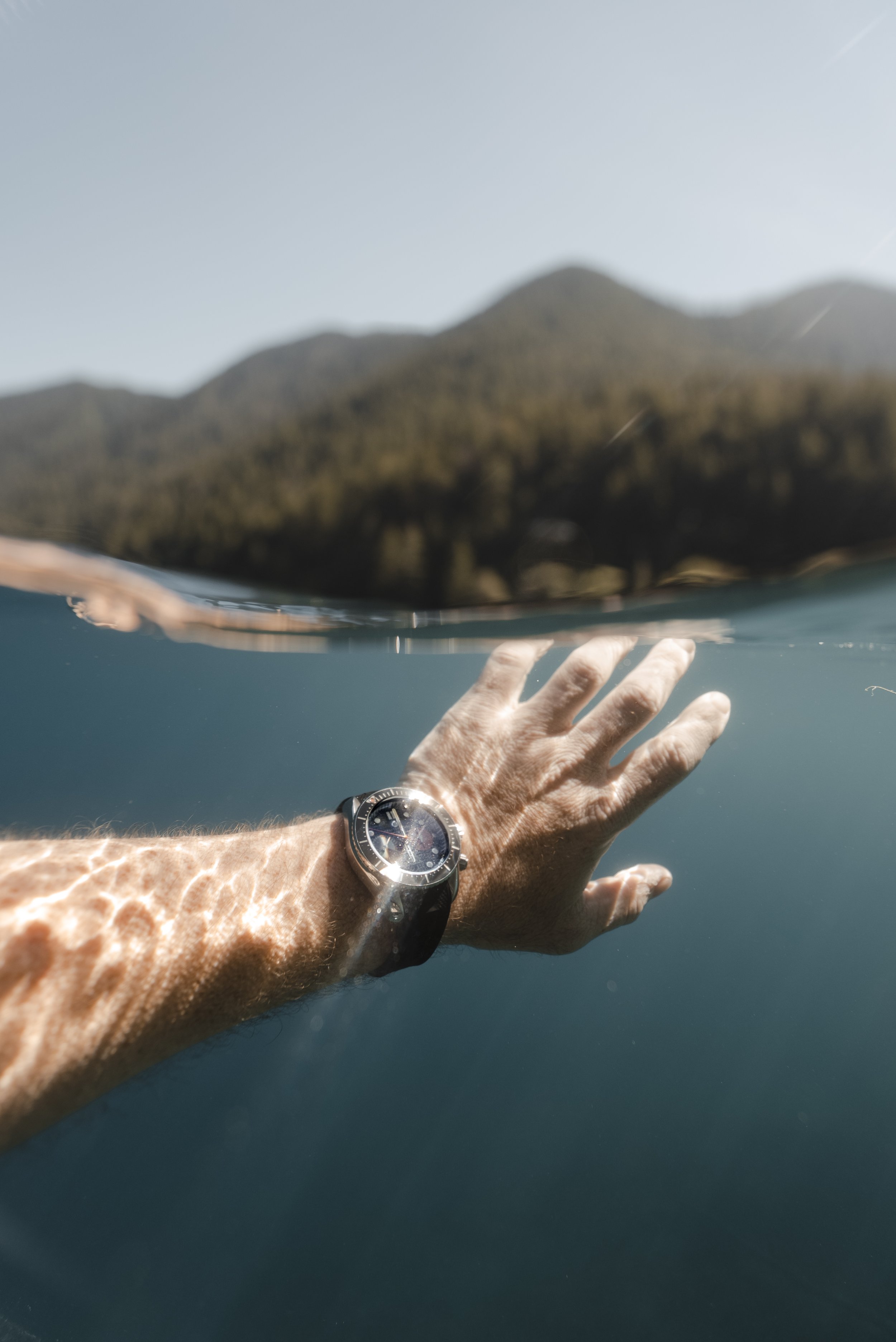 Over-under photo of a person's arm and hand reaching out of the water in a lake or ocean, wearing a Crafter Blue watch with a dark face, with mountains and a clear sky in the background.