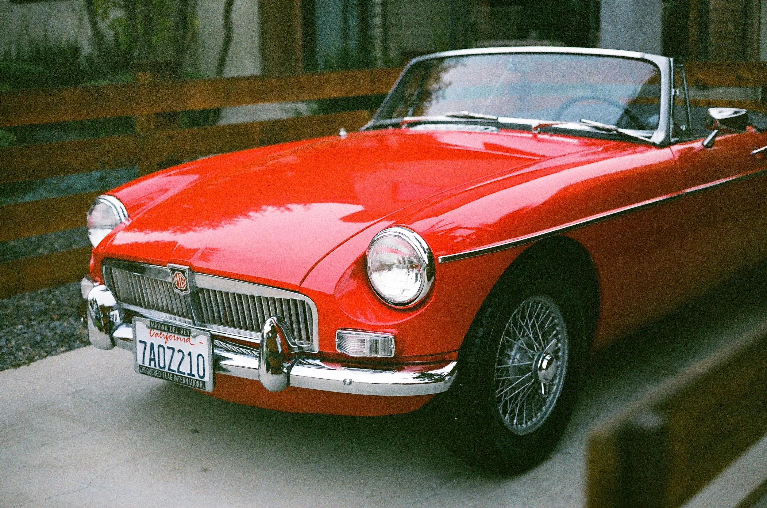 A classic red MG convertible sports car parked on a driveway, with a wooden fence and a house in the background.
