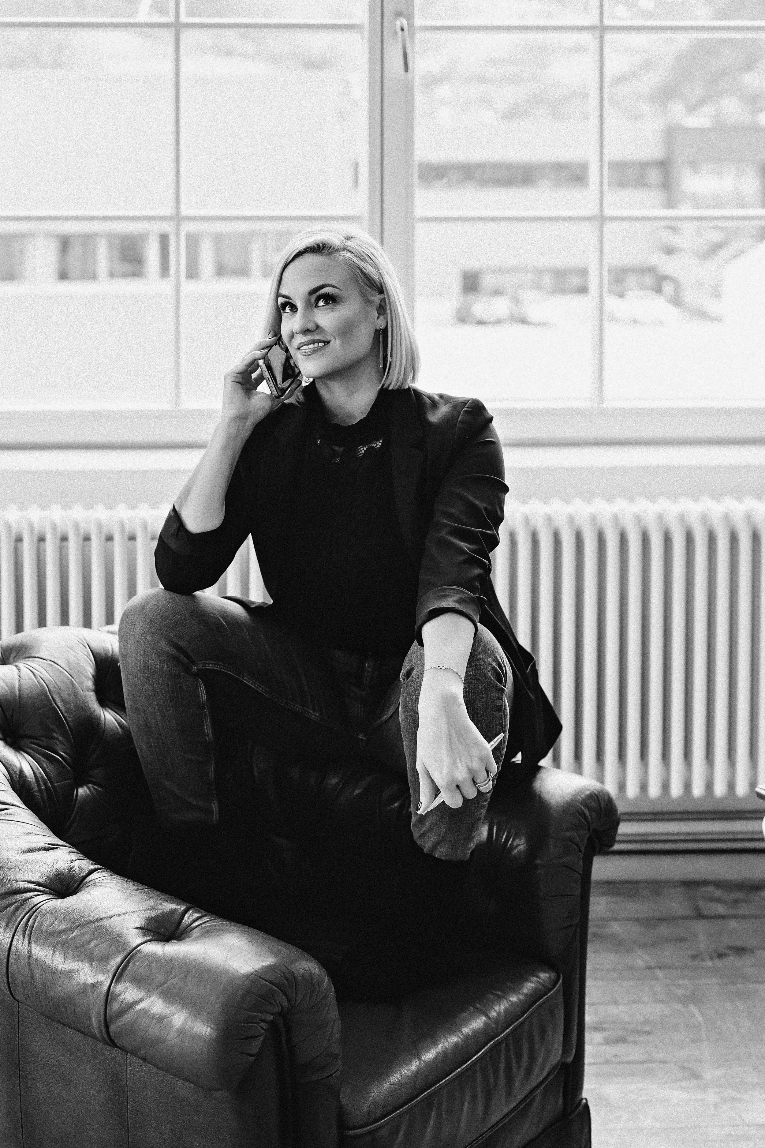 A woman sitting on a leather armchair, talking on a smartphone, with a large window behind her showing a cityscape.