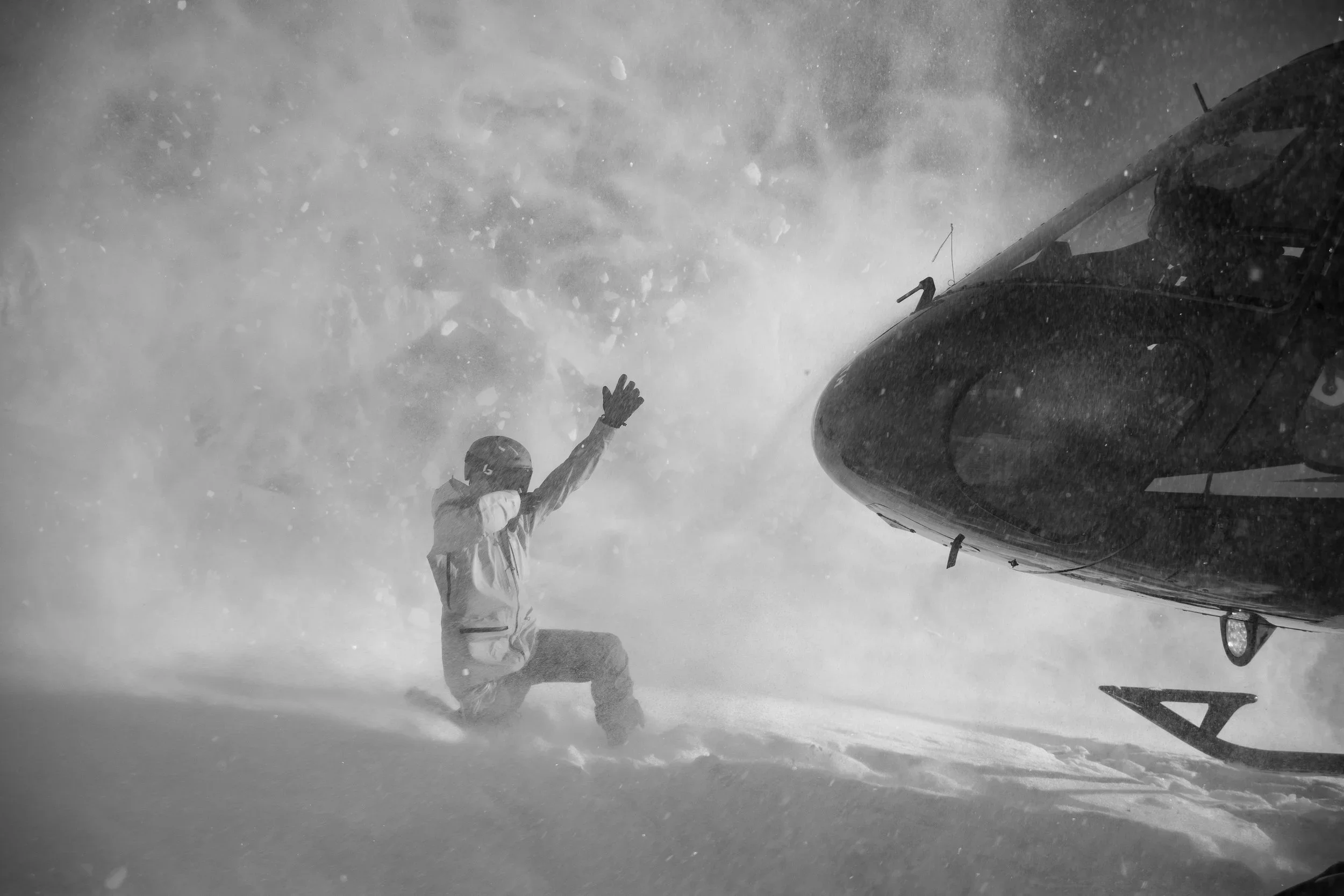 Points North Heli-Adventures staff kneeling in snow, guiding a helicopter hovering very close, with snow and wind flying around in a snowy environment.