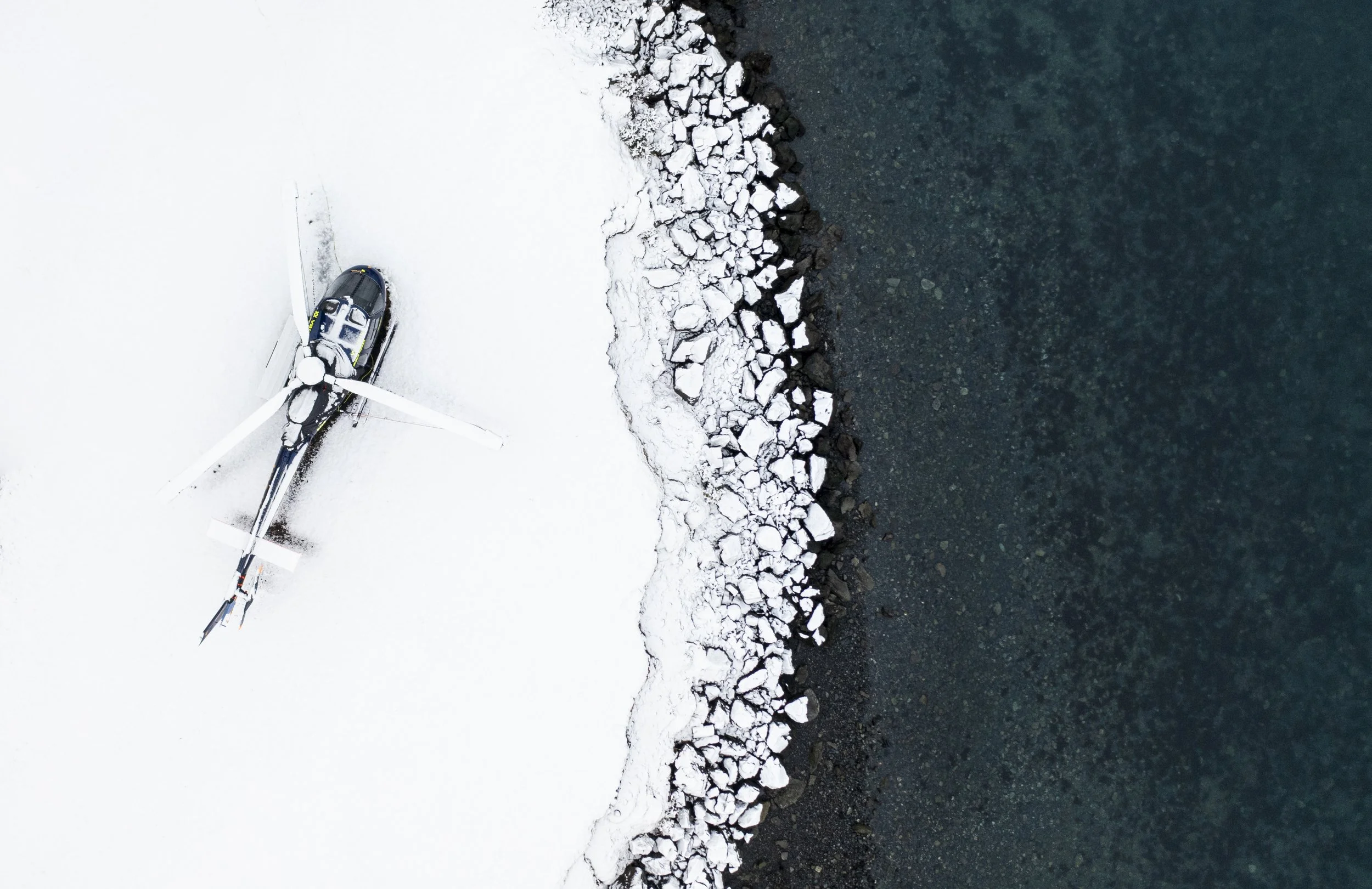 A helicopter resting on snow-covered ground near a rocky shoreline with dark waters.