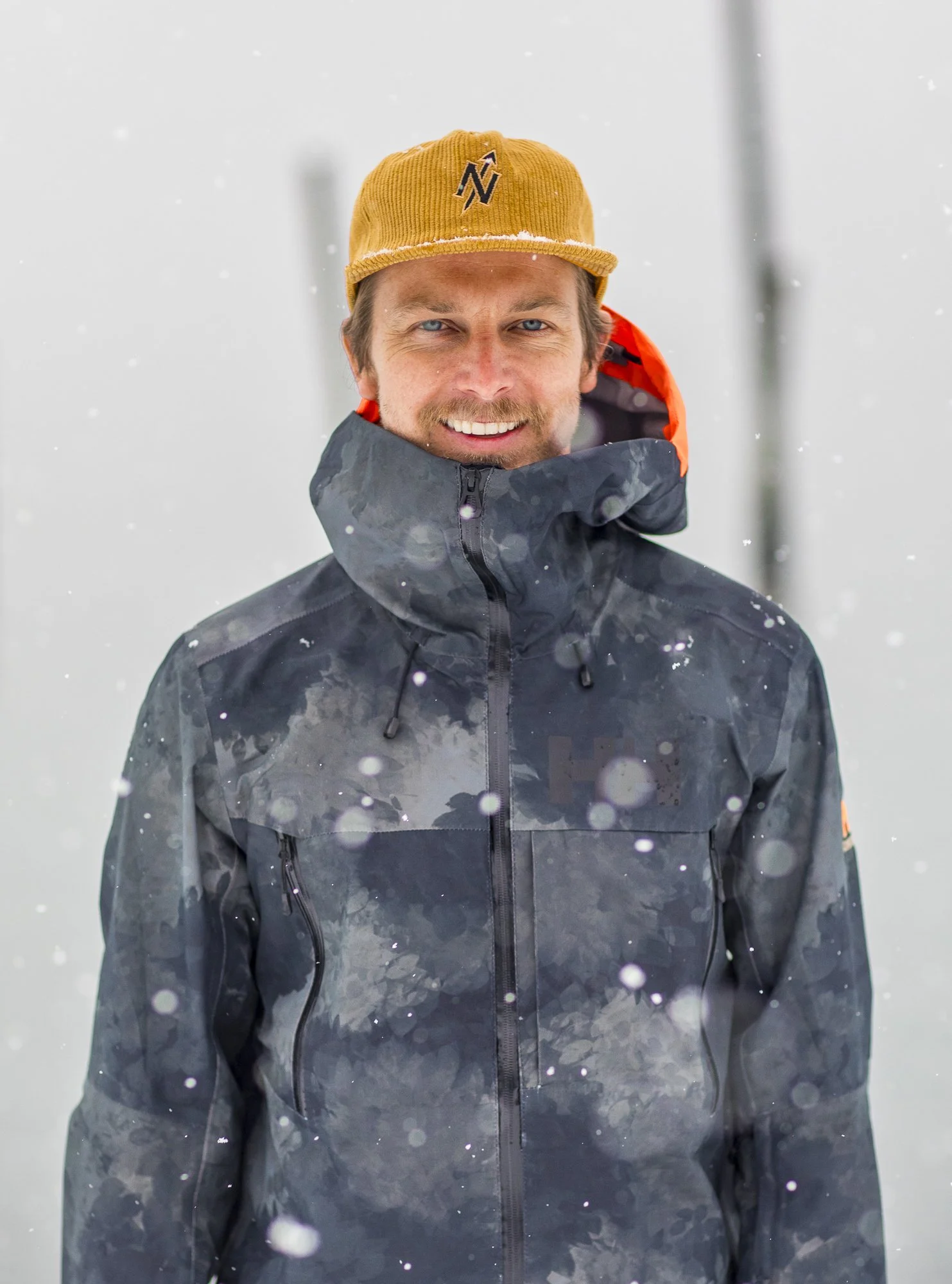 A man smiling outdoors in snowy weather, wearing a black waterproof jacket and a mustard yellow hat.
