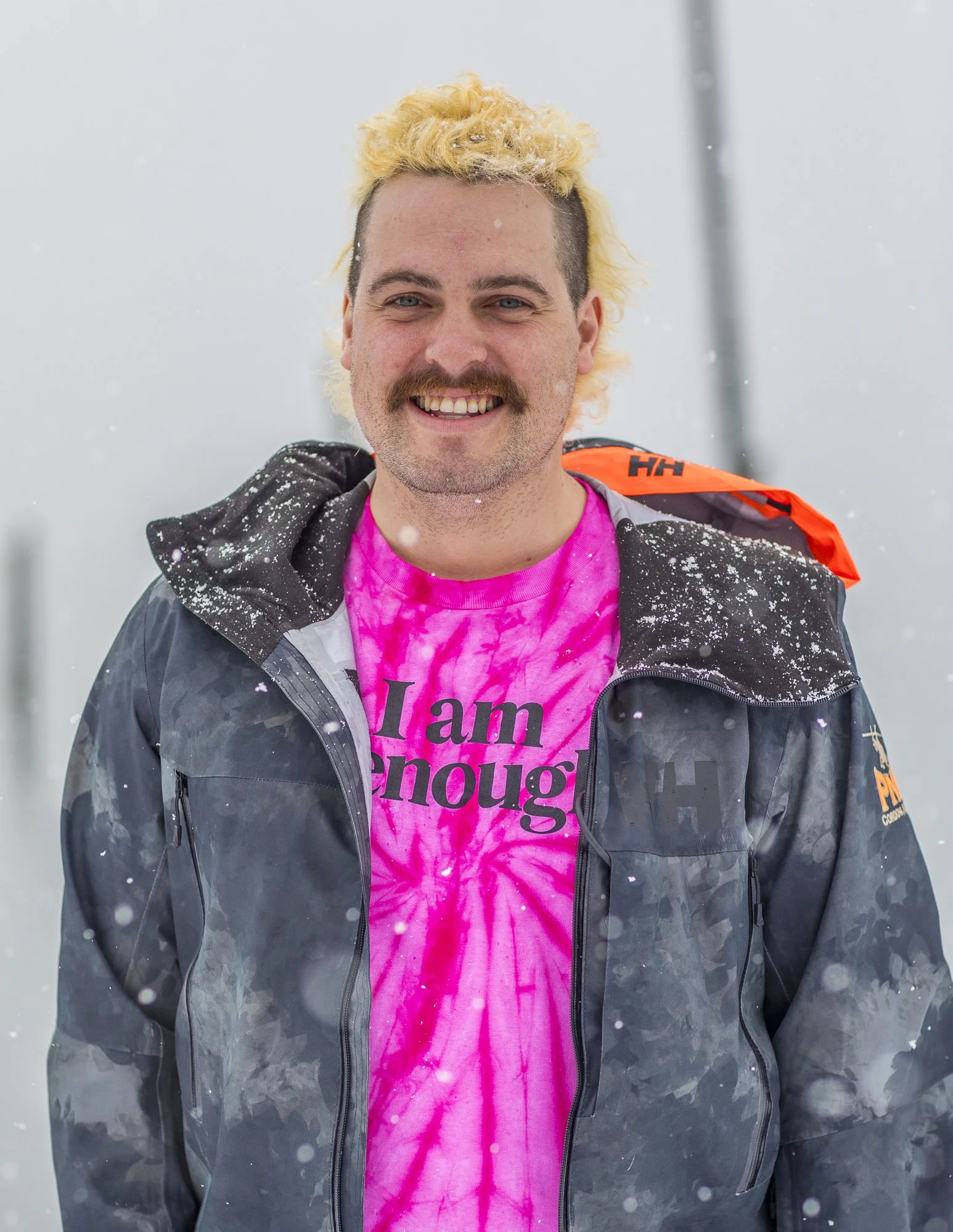 A smiling man with a blond, curly hairstyle and a mustache, wearing a bright pink tie-dye shirt with the words 'I am enough' and a dark jacket outdoors in snowy weather.