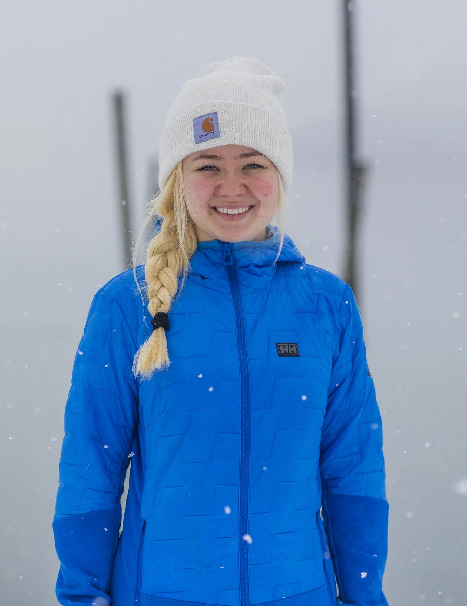 A young woman with a braid, wearing a white beanie and a blue jacket, smiling in a snowy outdoor setting.