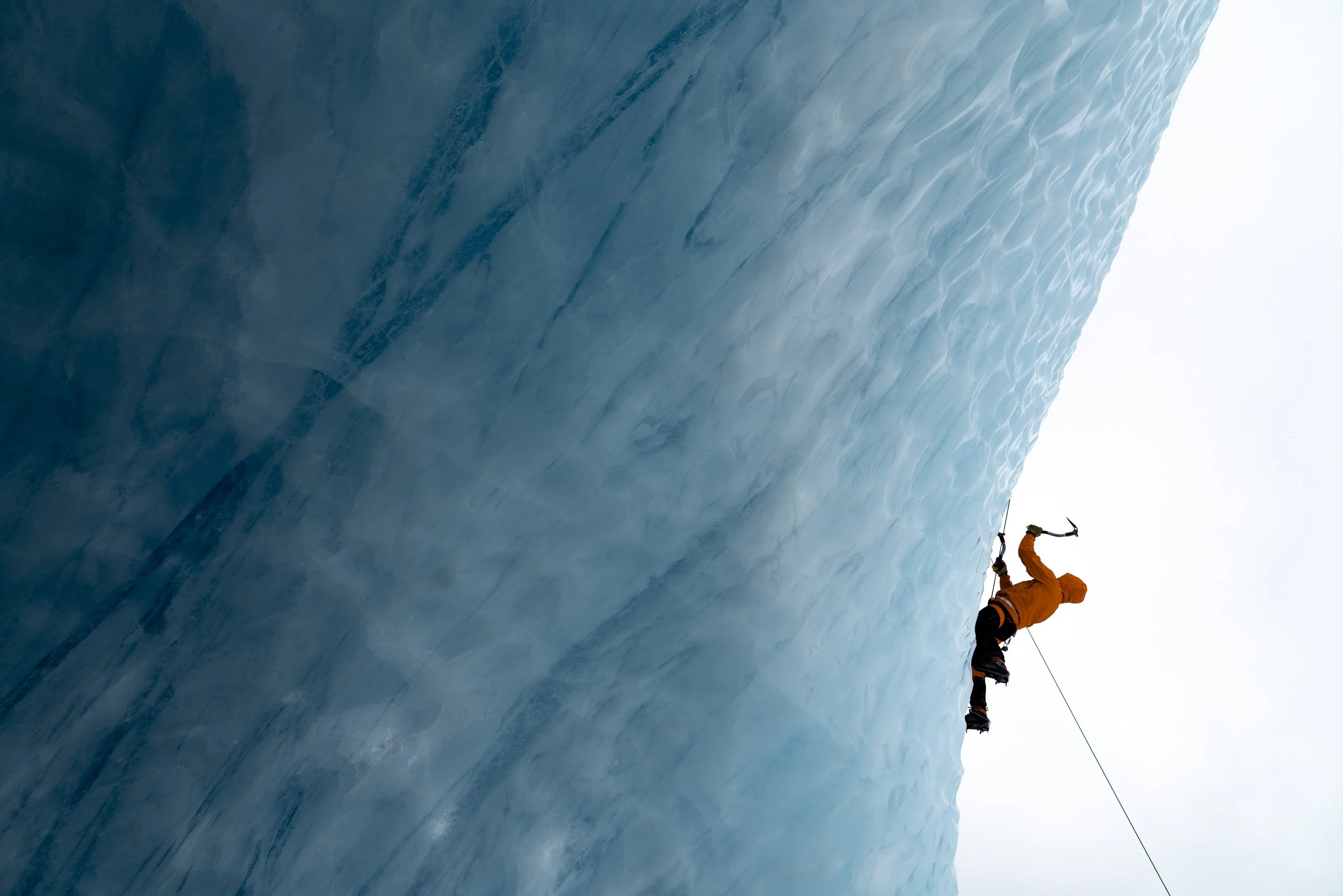 A person in orange climbing gear scaling a steep icy glacier, using an ice axe and harness for safety. This is an optional activity on PNH activity days.