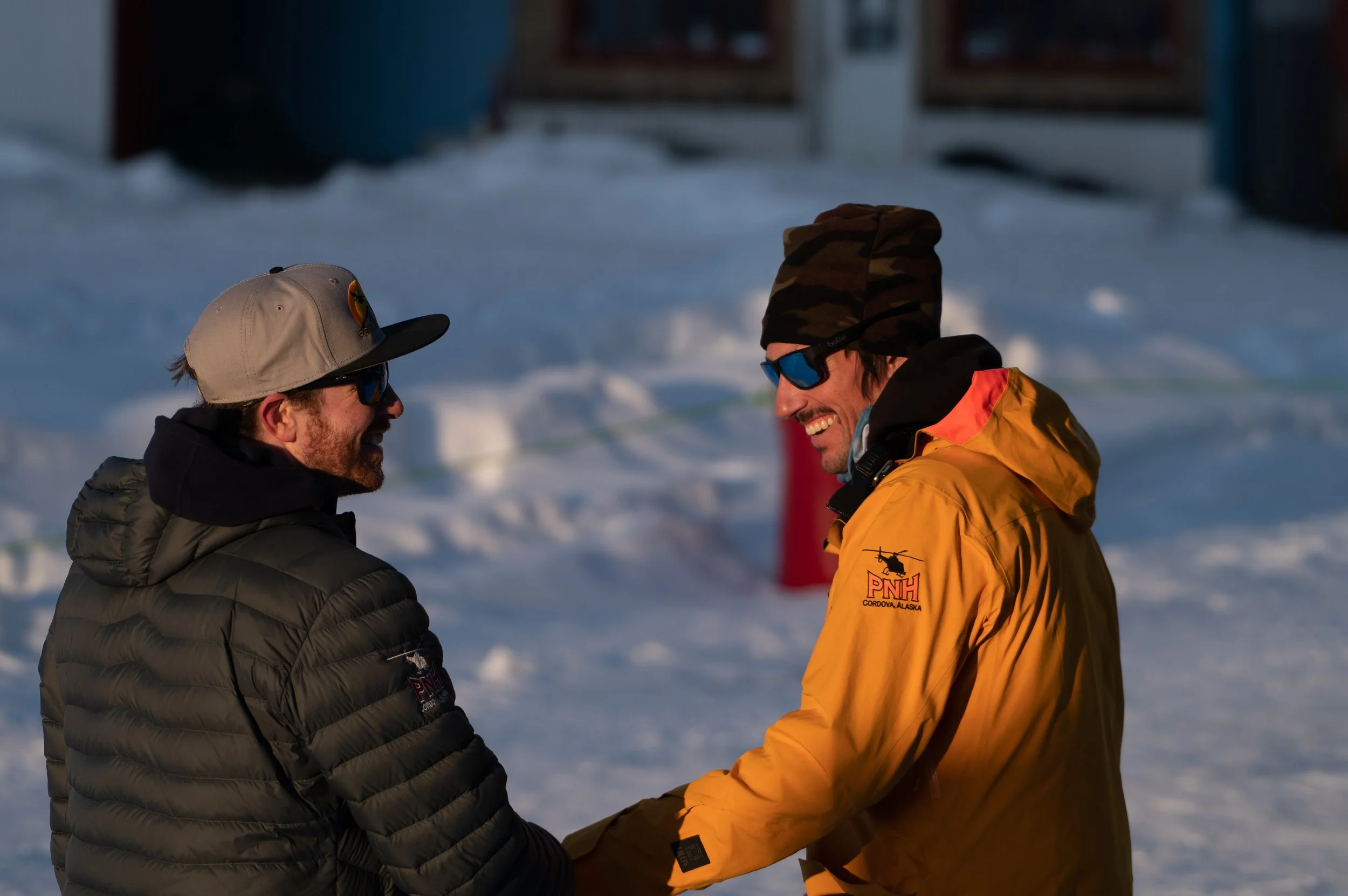 Two men outside in a snowy area, smiling and holding hands, wearing winter jackets, sunglasses, and hats.Clouds in the sky and a building are in the background.