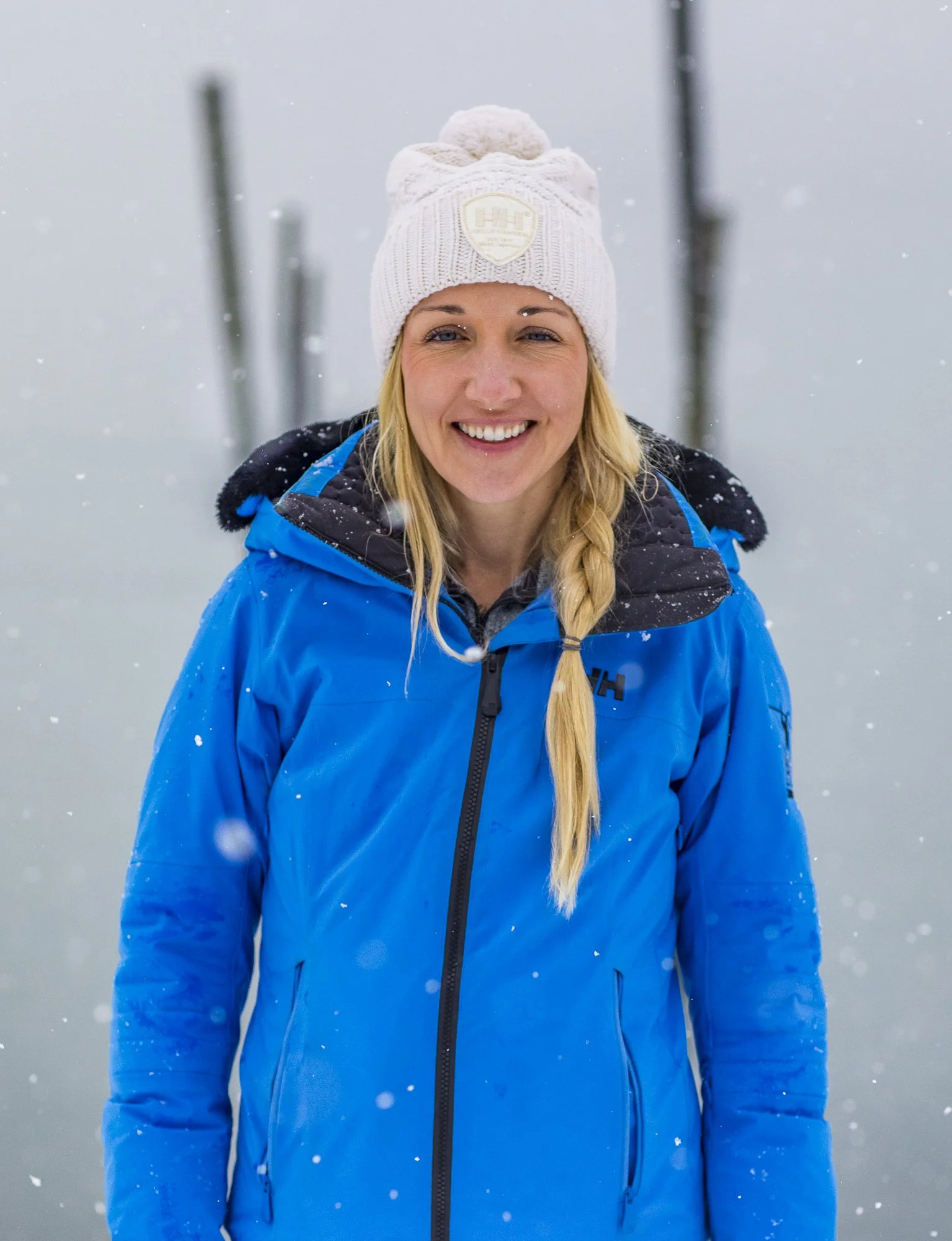 A smiling woman in a blue jacket and white knit hat standing in snowy outdoors.